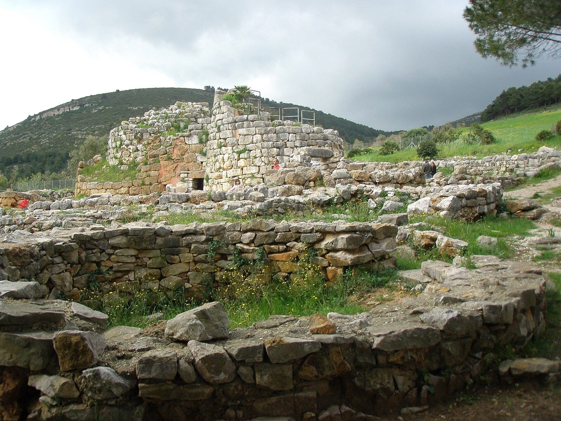 Ruinas de piedra de Nuraghe Palmavera con cabañas circulares y una torre central, frente a colinas verdes bajo un cielo nublado cerca de Alghero.