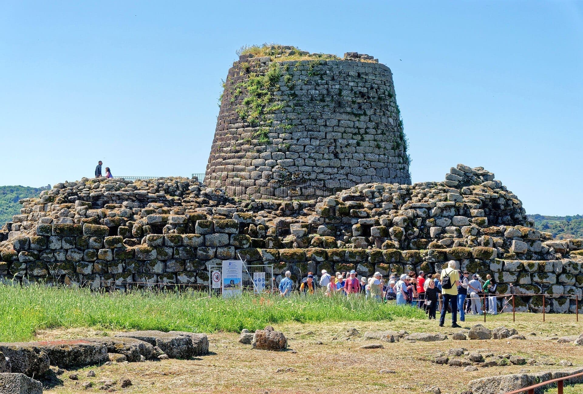 Las murallas de piedra y la torre central del Nuraghe Santu Antine se elevan sobre ruinas cubiertas de hierba, con grupos de visitantes explorando en un día soleado.