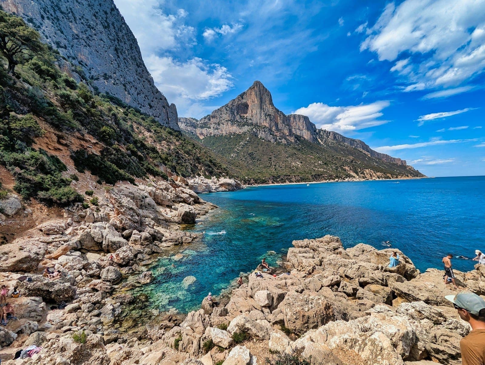 Imponentes acantilados de montaña sobre una costa rocosa de Cerdeña con aguas turquesas y un mar azul profundo bajo un cielo luminoso con nubes dispersas.