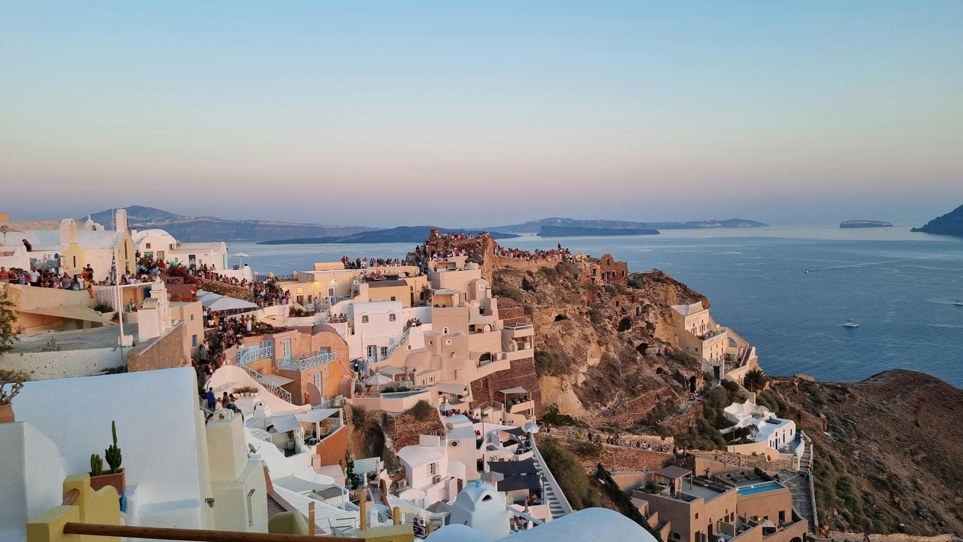 Weitblick auf den Sonnenuntergangs-Aussichtspunkt in Oia mit Hunderten von Besuchern an den Burgruinen, weißen Häusern und der Ägäis während der goldenen Stunde.
