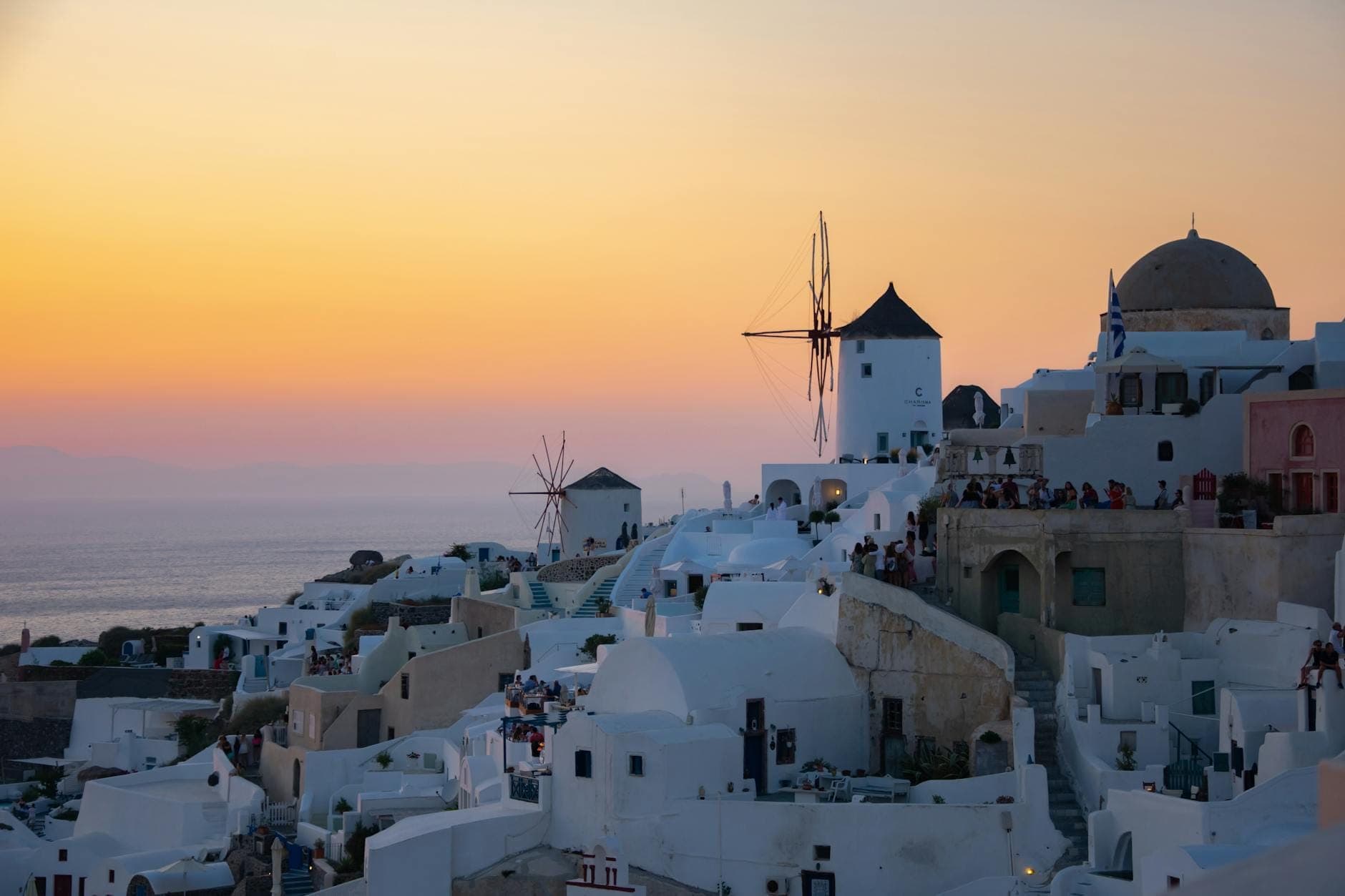 Die Windmühlen von Oia auf Santorini bei Sonnenuntergang, hoch über den charakteristischen weißen Häusern, während sich Besucher versammeln und der Himmel in goldenen und orangefarbenen Tönen über der Ägäis leuchtet.