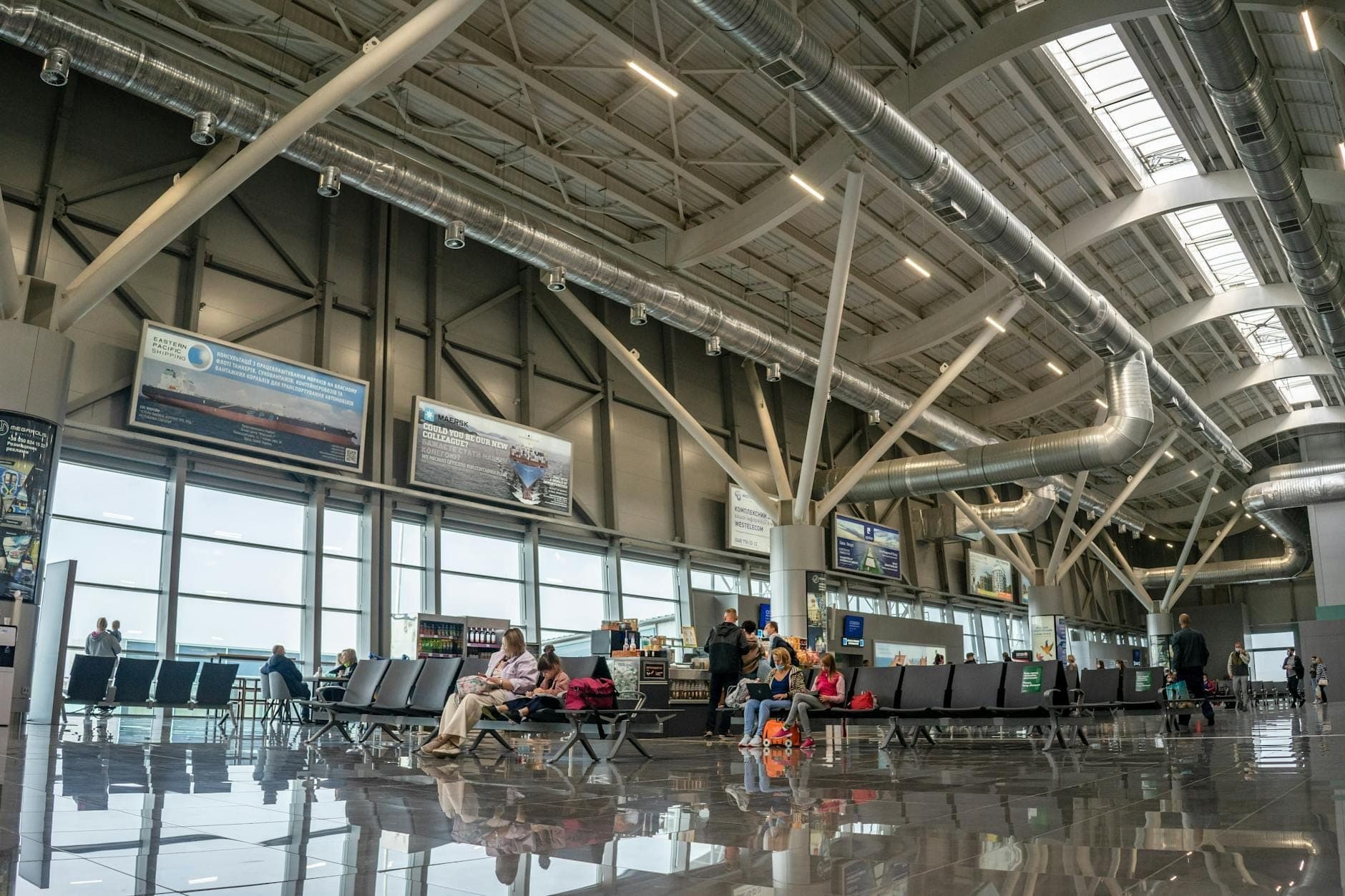 Interior moderno de terminal de aeropuerto con pasajeros sentados en filas de asientos, grandes ventanas y techos altos con vigas vistas.