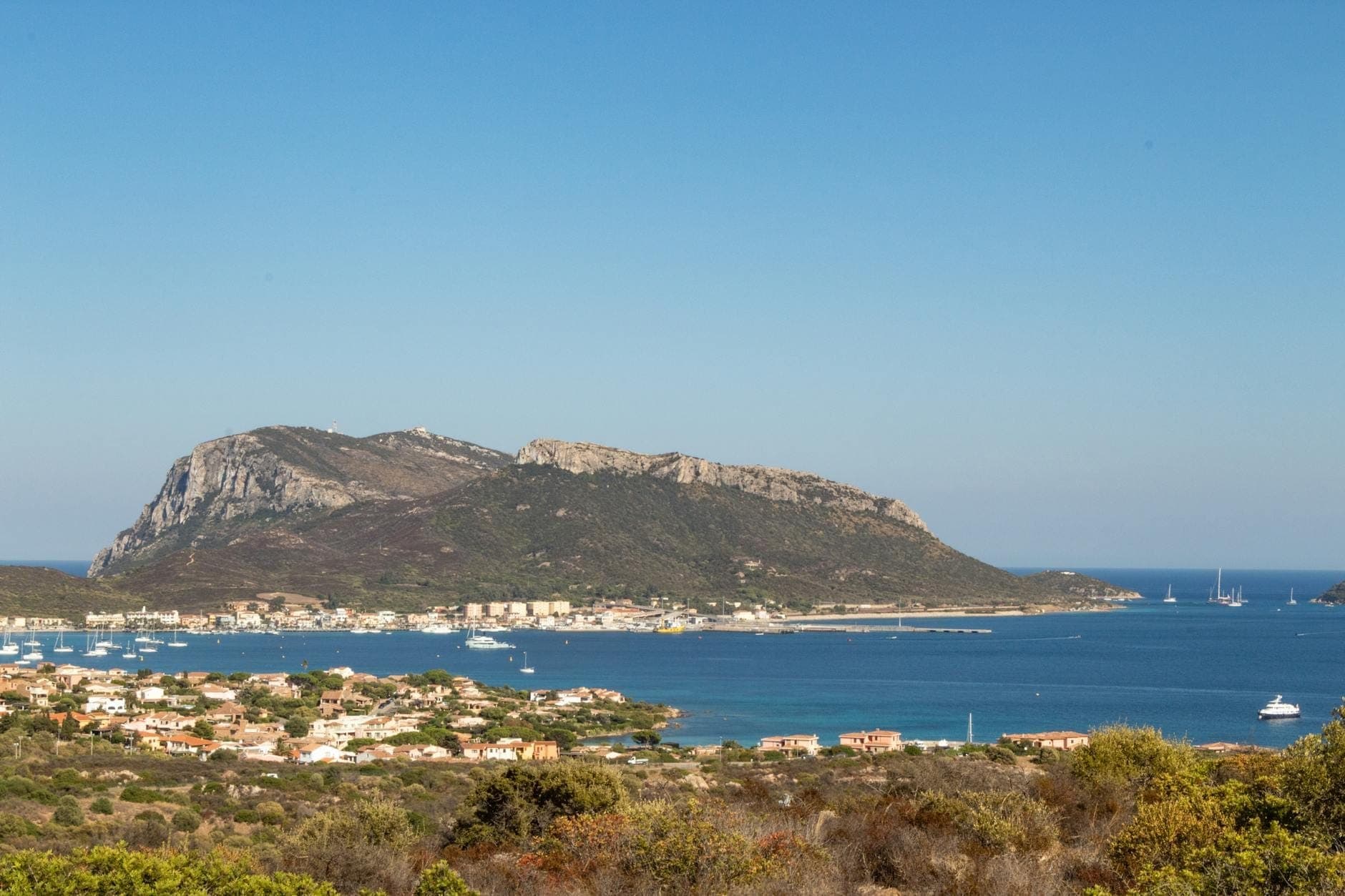 Vista panorámica de Olbia con el mar, veleros, la ciudad costera y una colina destacada al fondo bajo un cielo azul despejado.