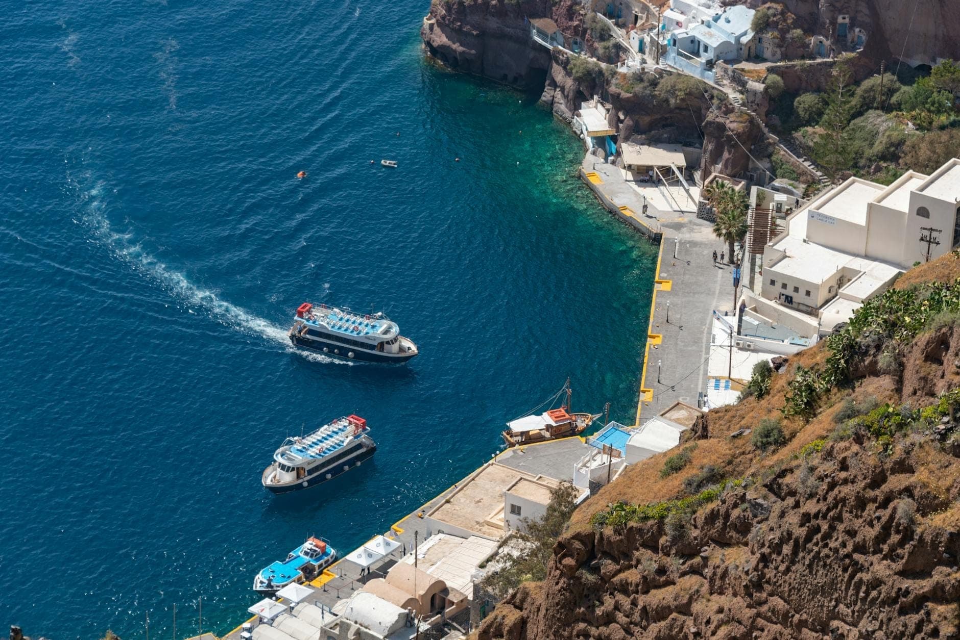 Luftaufnahme des alten Hafens von Fira auf Santorini mit zwei angedockten Booten, vor den beeindruckenden vulkanischen Caldera-Klippen und dem blauen Meer.