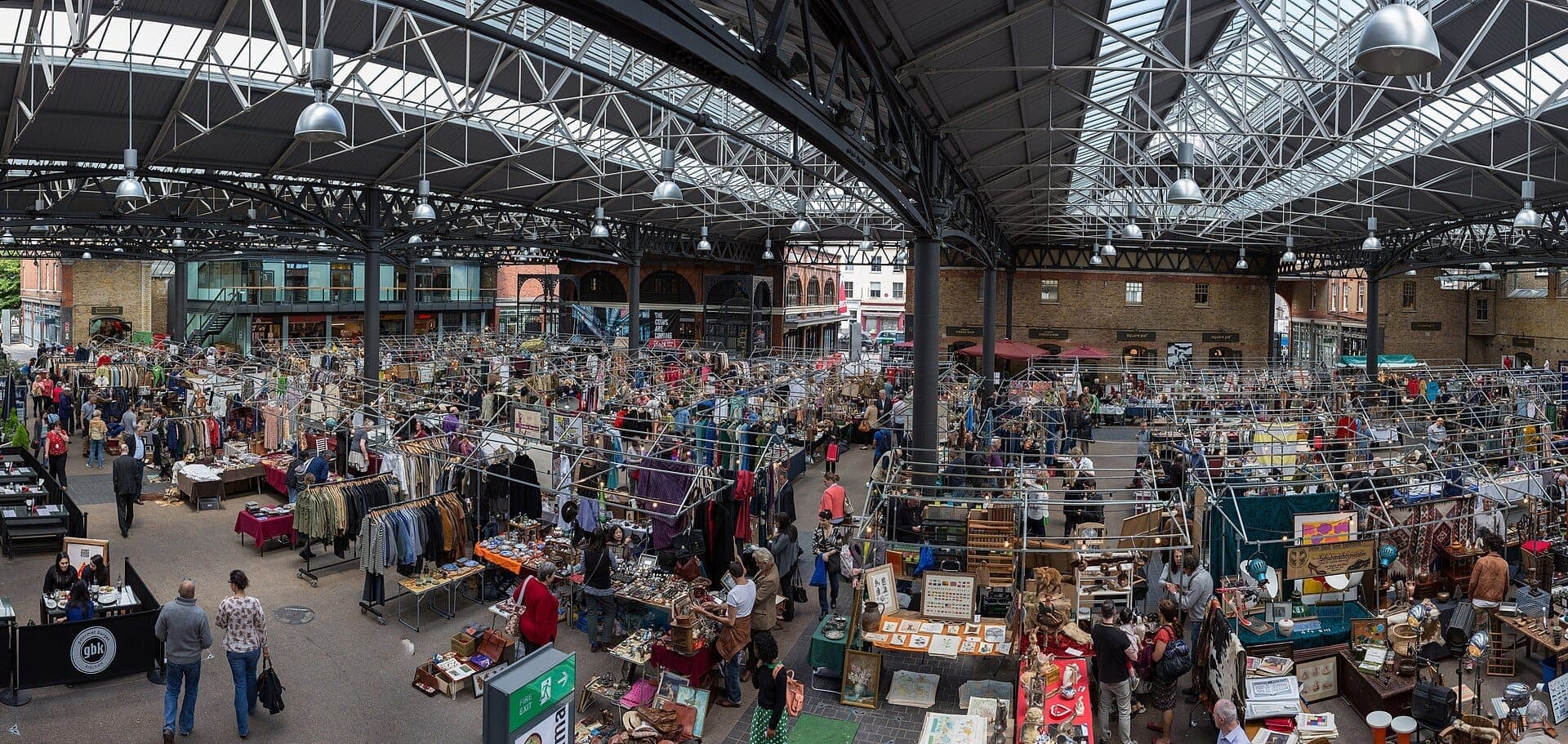 Weitwinkelansicht des Innenraums des Old Spitalfields Market mit belebten Menschenmassen, Händlern, Vintage-Kleidung und Waren unter einem Eisen- und Glasdach.