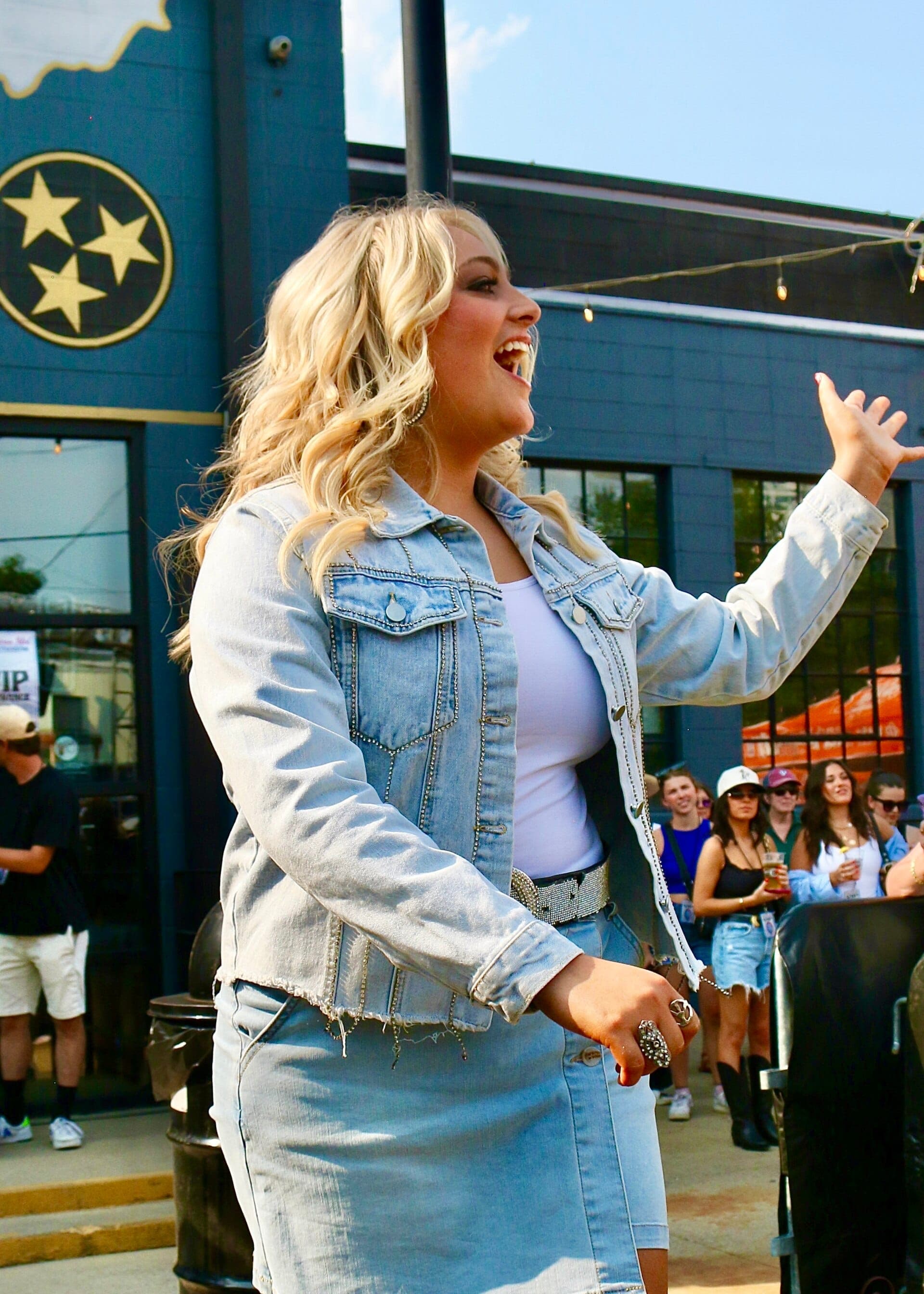 A woman in a denim jacket smiles and gestures outside Ole Smoky Distillery at 6th & Peabody, with people gathered behind her.