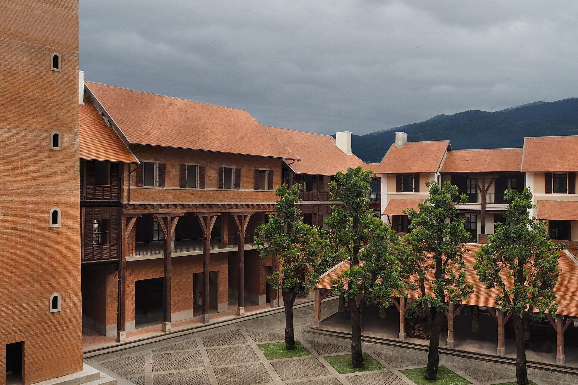 Edificios de ladrillo con tejados de terracota rodean una tranquila plaza flanqueada de árboles verdes en One Nimman en Chiang Mai, bajo un cielo nublado.