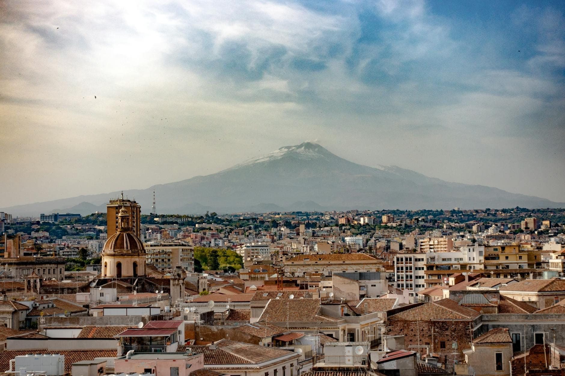 Panoramikong tanawin ng lungsod ng Catania na may niyebeng tuktok ng Monte Etna na bantayog sa likod sa ilalim ng dramatikong langit.