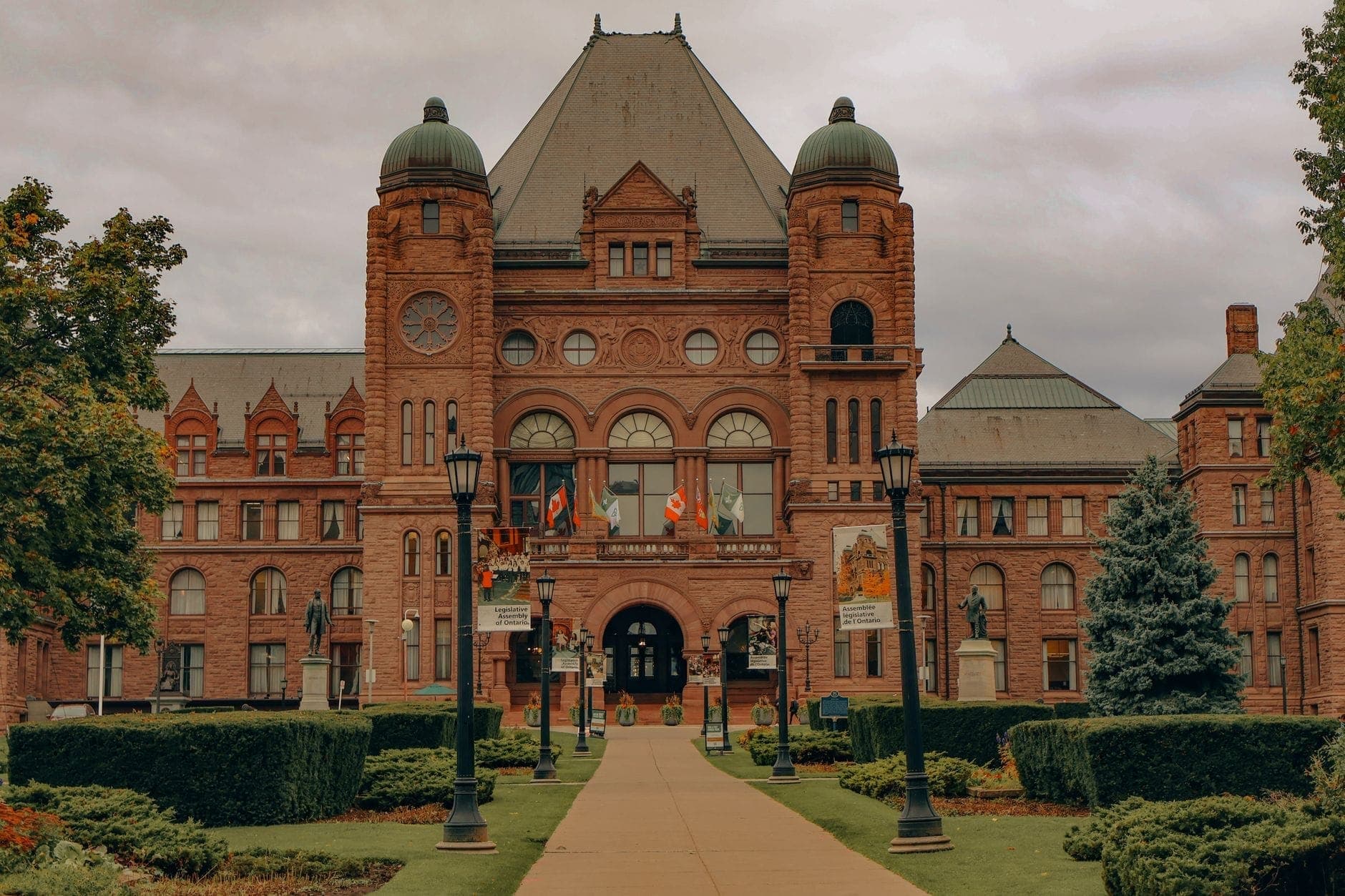 L'Edificio del Parlamento dell'Ontario a Toronto, un imponente edificio in arenaria con cupole verdi e giardini curati, visto dal vialetto principale d'ingresso in una giornata nuvolosa.