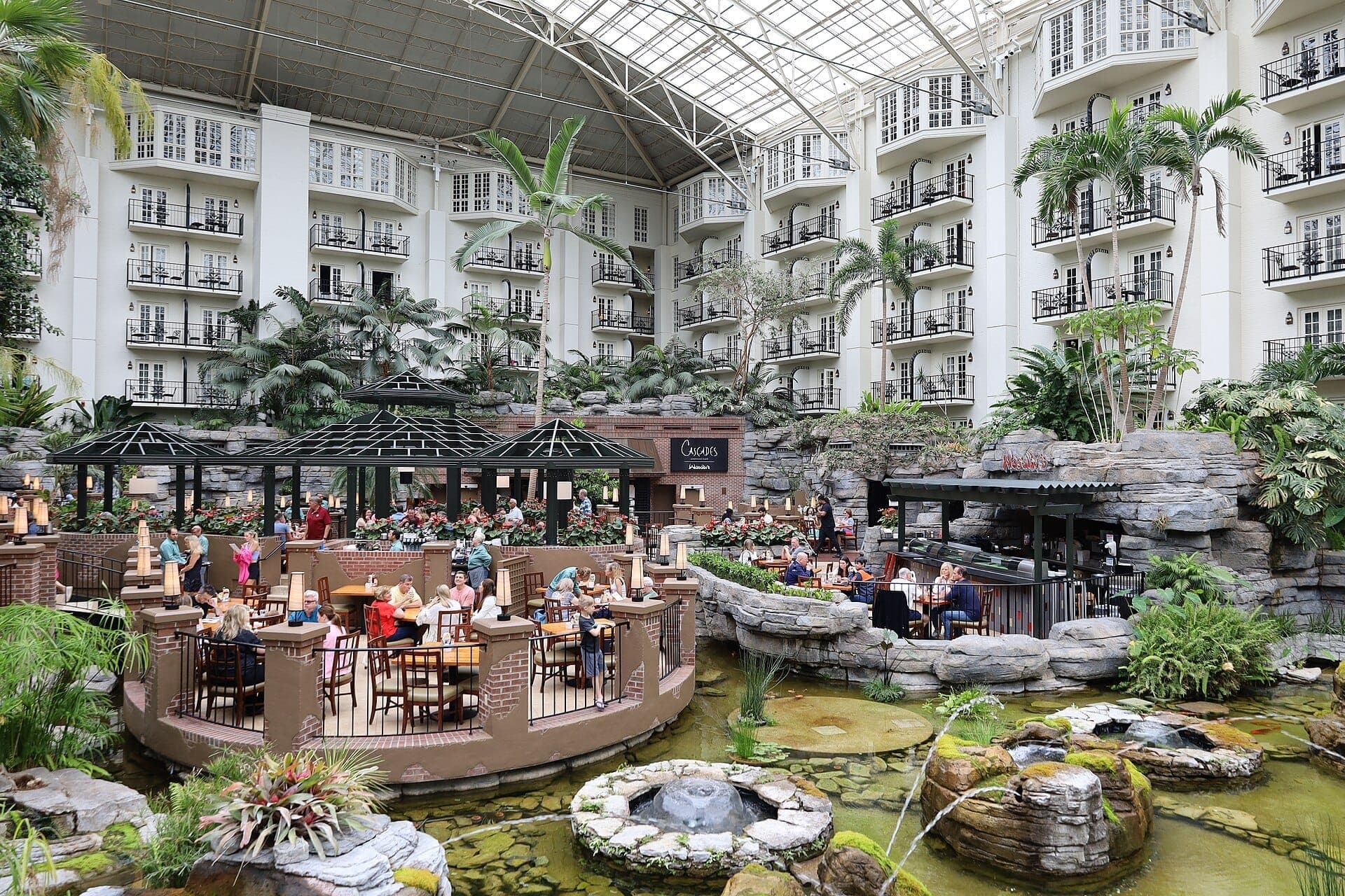 Spacious atrium at Gaylord Opryland Resort with dining patios, lush tropical plants, water features, and hotel balconies under a glass roof.