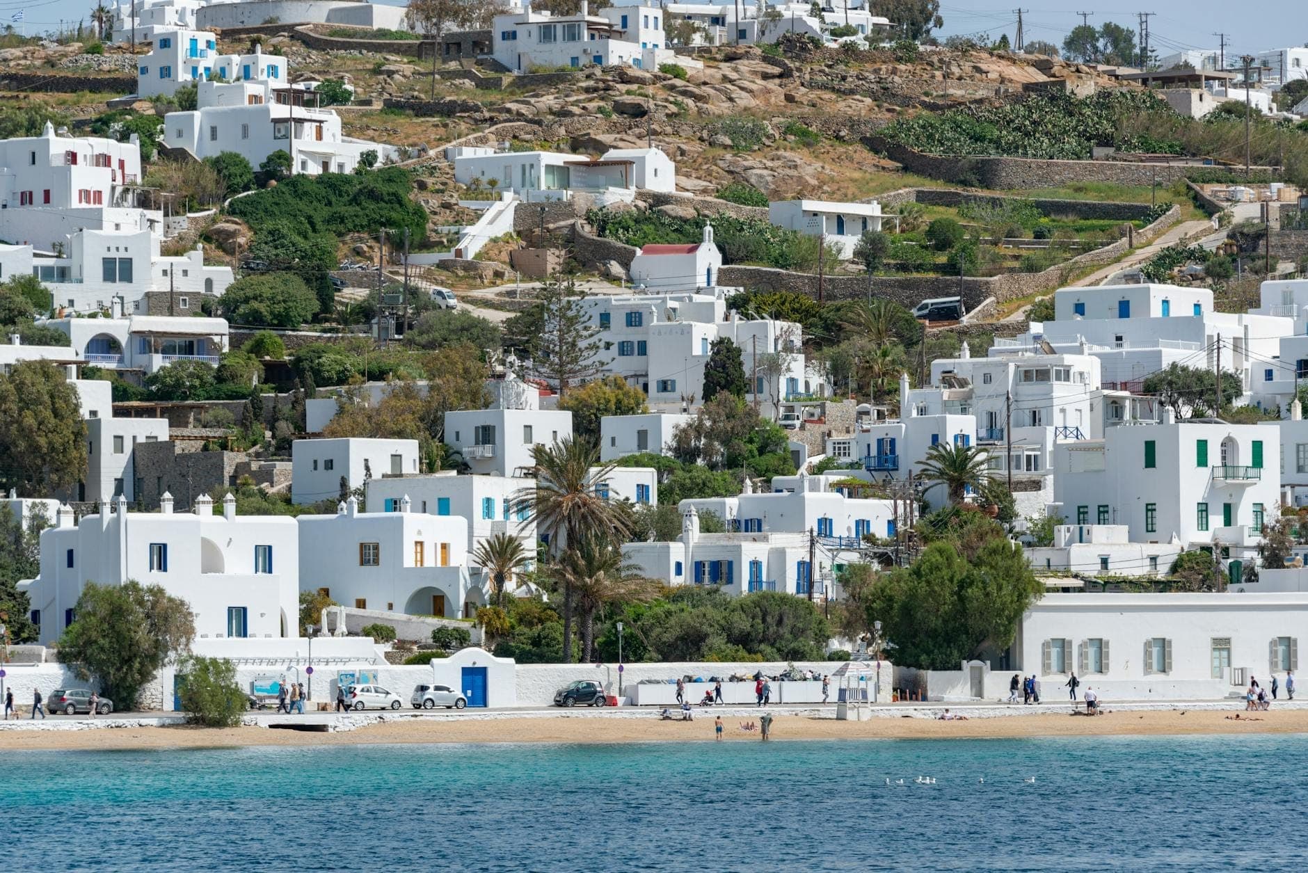 Weitläufiger Blick auf den Ornos Beach mit Menschen am Sandstrand, klarem türkisblauen Wasser und weißen kykladischen Häusern, die sich den Hügel hinaufziehen.