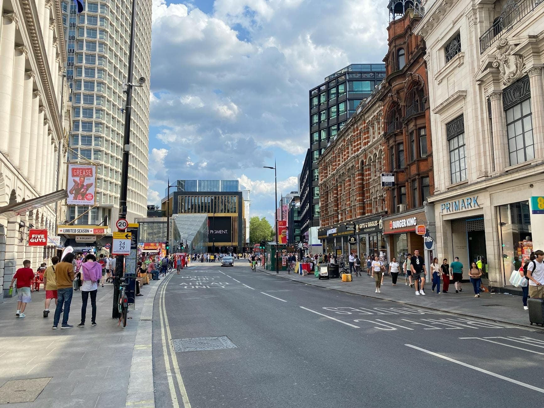 Lebhafter Blick auf die Oxford Street tagsüber mit Einkaufenden auf den Bürgersteigen, Flagship- und Modekettenstores, klassischer und moderner Londoner Architektur sowie einem teils bewölkten Himmel.