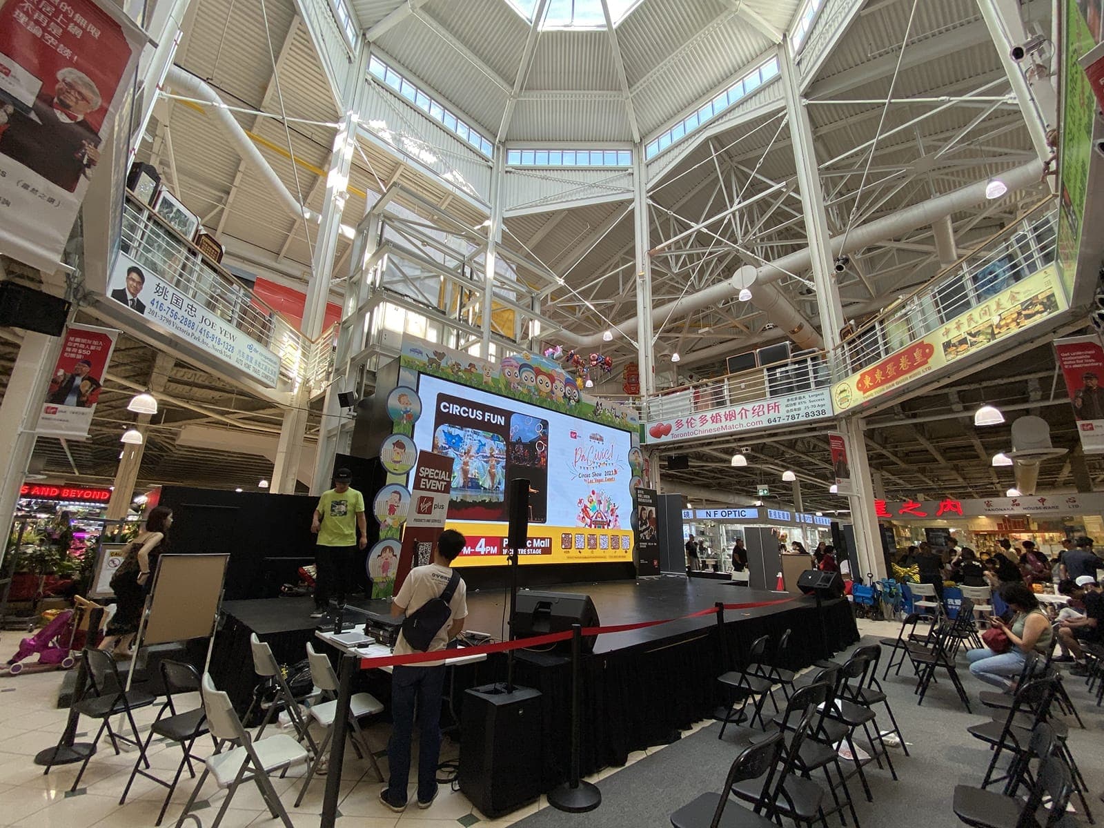 Vista amplia del atrio central iluminado de Pacific Mall, con un escenario, pantalla digital de eventos, sillas y personas bajo un techo con claraboya.