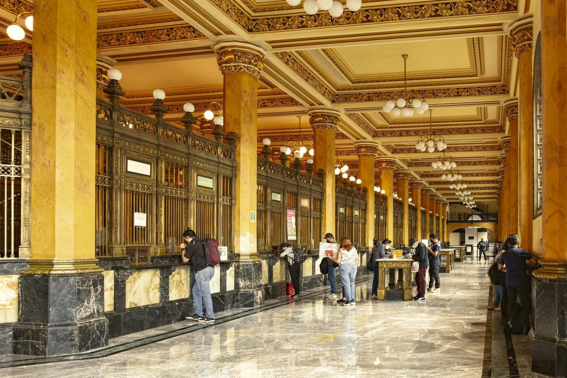 ผู้คนติดต่อเคาน์เตอร์หินอ่อนและทองสุดหรูภายใน Palacio Postal ของเม็กซิโกซิตี้ รายล้อมด้วยเสาสีทองสุดอลังการและเพดานตกแต่งอย่างละเอียดอ่อน