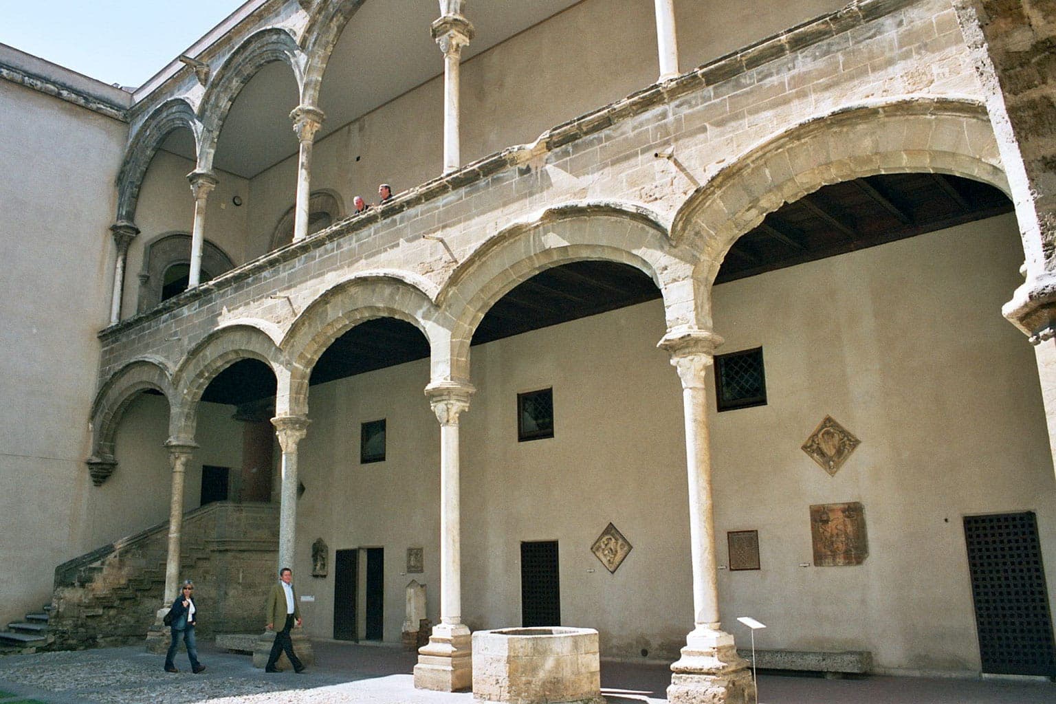 Halaman dalam Palazzo Abatellis dengan lengkungan batu yang elegan, galeri atas, dan pengunjung yang menjelajahi arsitektur Gotik-Katalan bersejarah di Palermo.