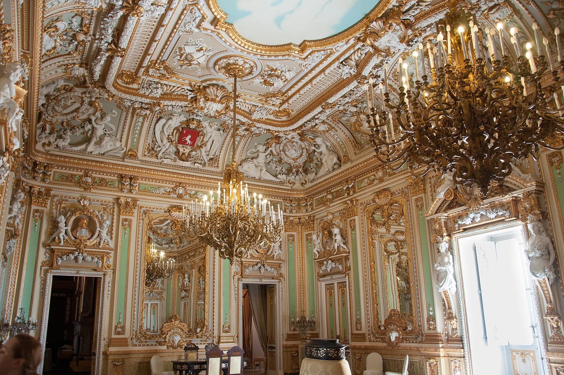 Prunkvoller vergoldeter Ballsaal im Palazzo Parisio mit barockem Stuckwerk, goldenen Zierleisten, großen Kronleuchtern und pastellfarbenen Wänden mit klassischen Statuen.