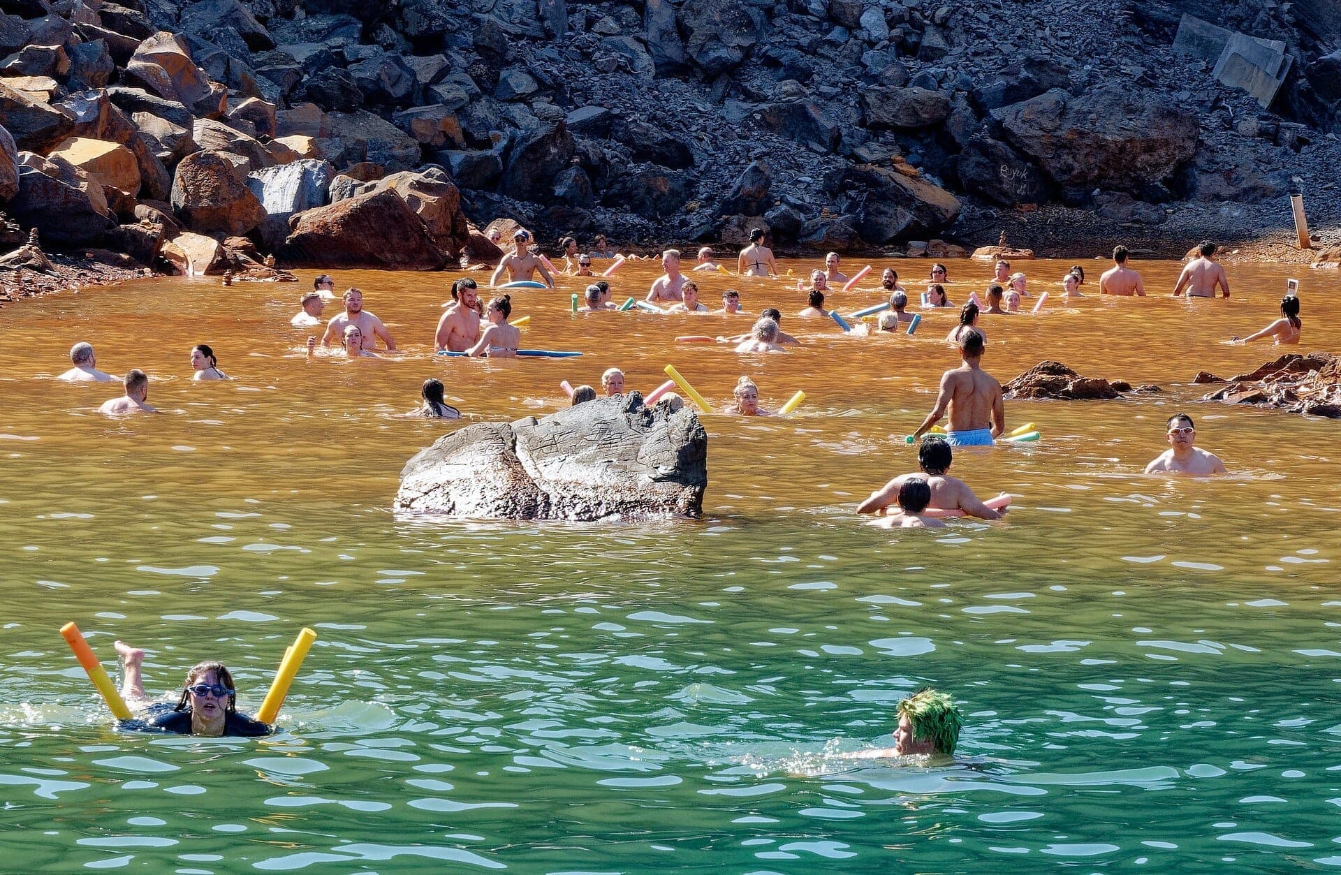 Dutzende Menschen schwimmen in den warmen, rostfarbenen Gewässern der Palea Kameni Hot Springs, umgeben von felsigem Vulkangelände.