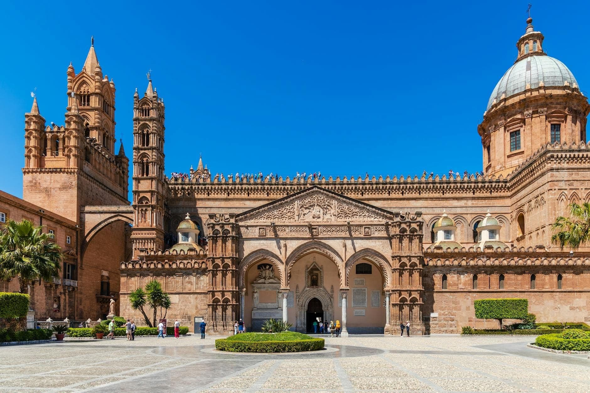 Pemandangan luas dan cerah fasad Katedral Palermo yang megah, dengan menara, pintu masuk beratap lengkung, dan kubah di bawah langit biru jernih, serta pengunjung yang berkeliling di alun-alun yang luas.