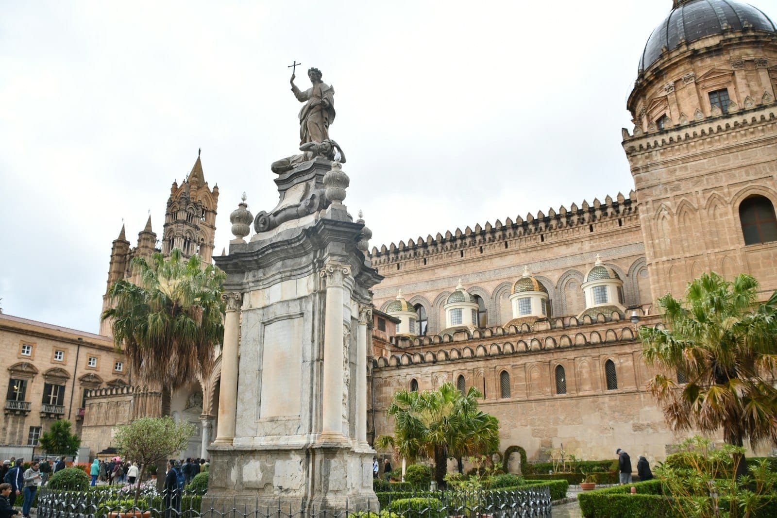 Patung dan pohon palem di depan Katedral Palermo yang megah dengan orang-orang berjalan di alun-alun pada hari berawan cerah.