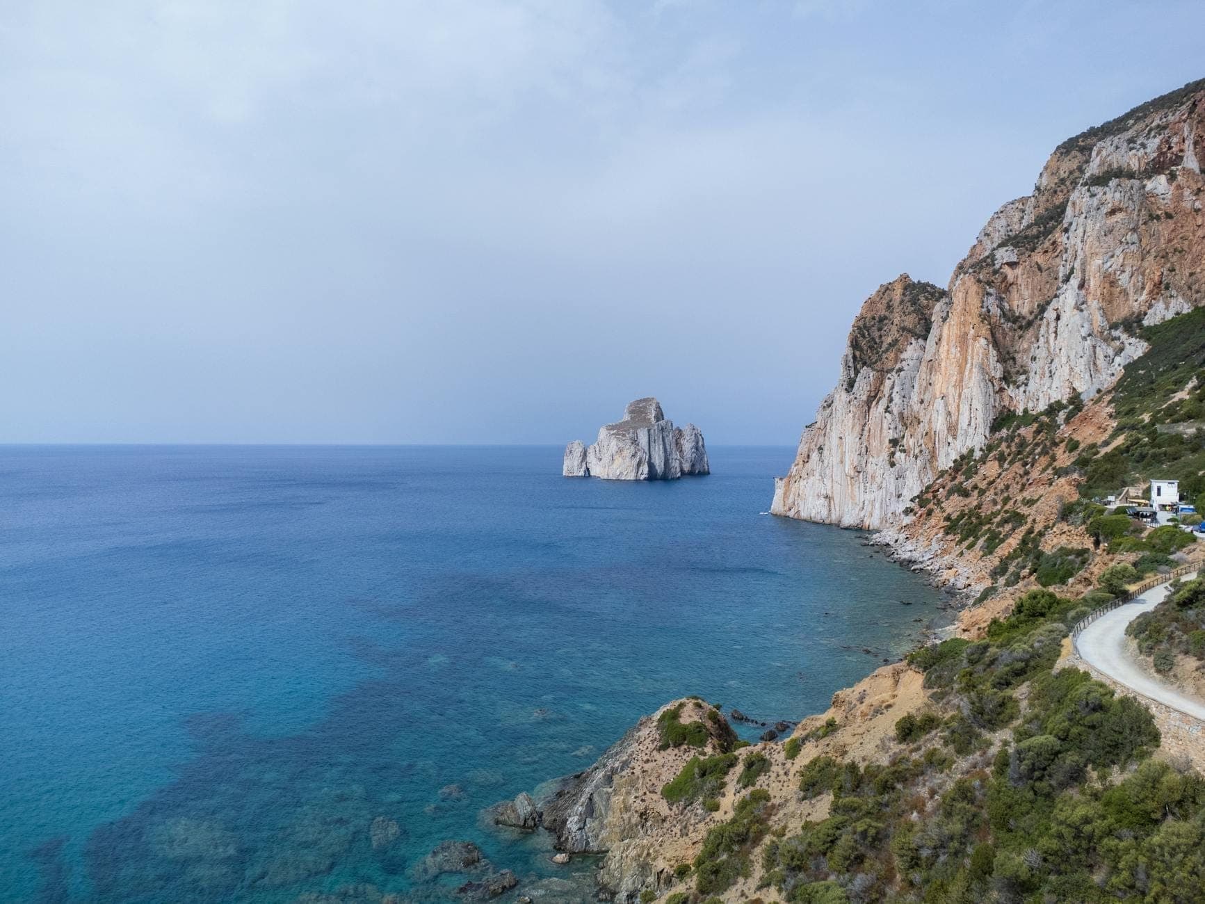 Vista panorámica del faraglione Pan di Zucchero emergiendo de las aguas turquesas junto a imponentes acantilados rocosos en la costa suroeste de Cerdeña, bajo un cielo despejado y luminoso.