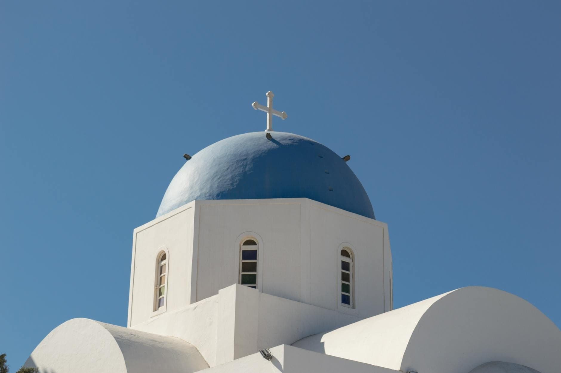 Weiße griechisch-orthodoxe Kirche mit blauer Kuppel und Kreuz, vor klarem blauem Himmel in Oia, Santorin, Griechenland.