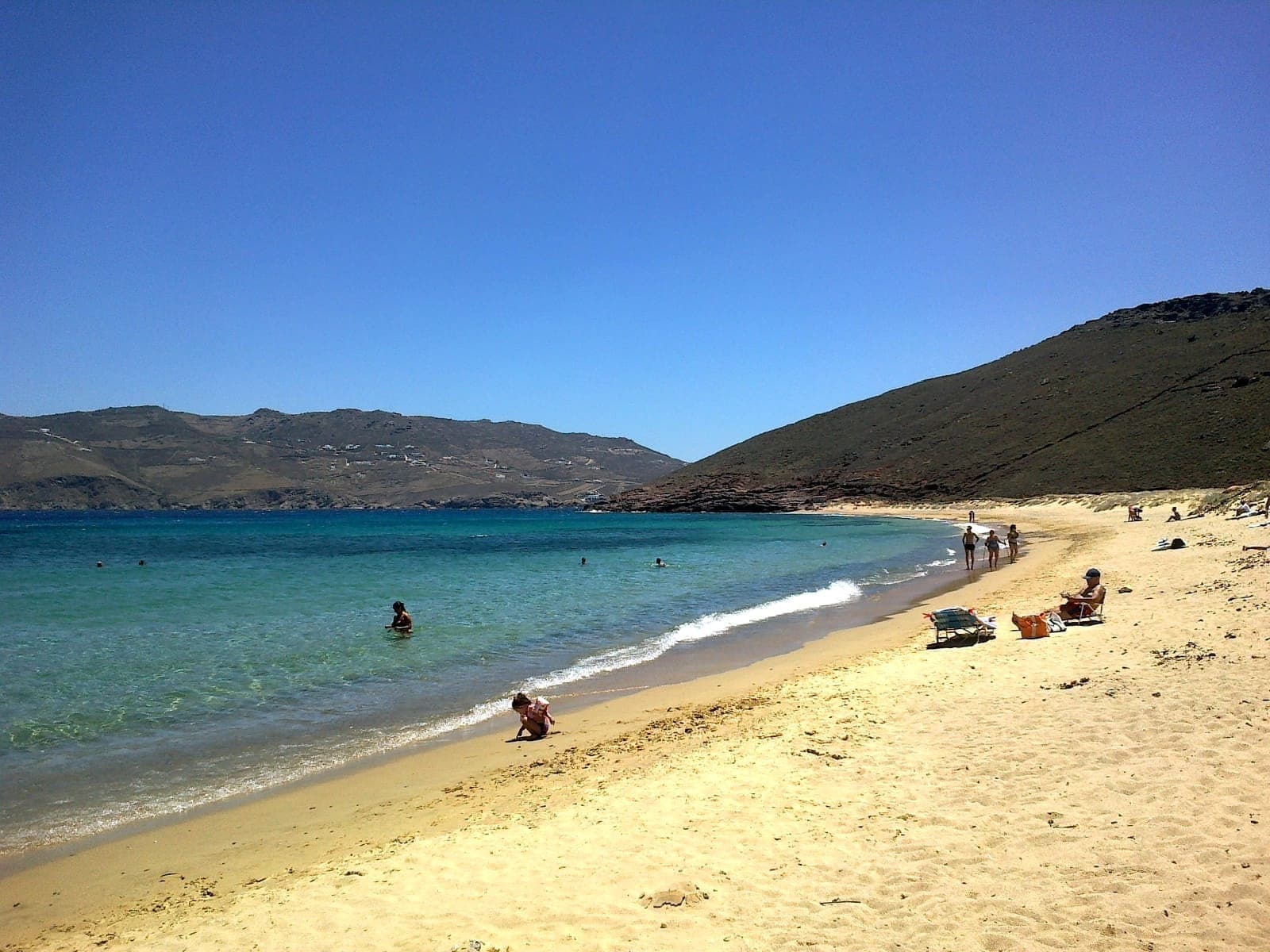 Goldener Sand und klares blaues Wasser verlaufen geschwungen entlang des Panormos Beach, mit einigen Sonnenbadenden und Schwimmern vor einer Kulisse felsiger Hügel.