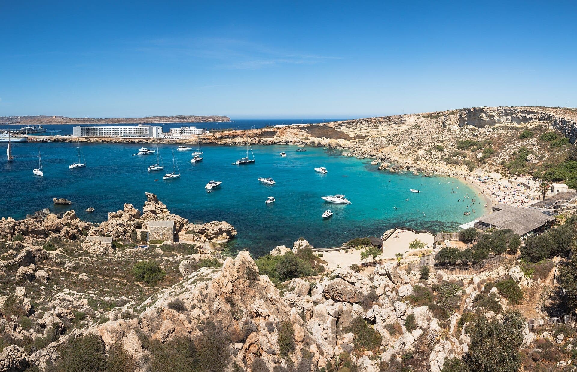Panoramablick auf Paradise Bay, Malta, mit türkisfarbenem Wasser, vor Anker liegenden Booten, felsiger Küste, Sandstrand und umliegenden Gebäuden unter klarem Himmel.