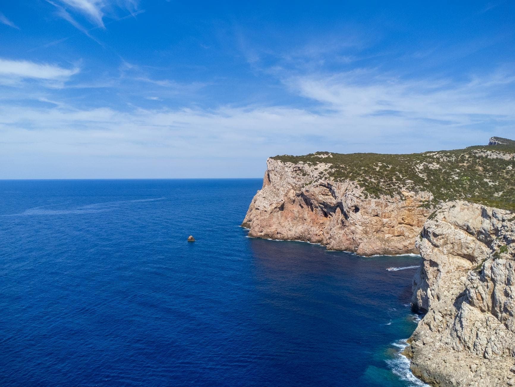 Dramatische Kalksteinklippen ragen unter blauem Himmel aus dem tiefen Blau des Meeres — ein Blick auf die raue Küste des Naturparks Porto Conte.