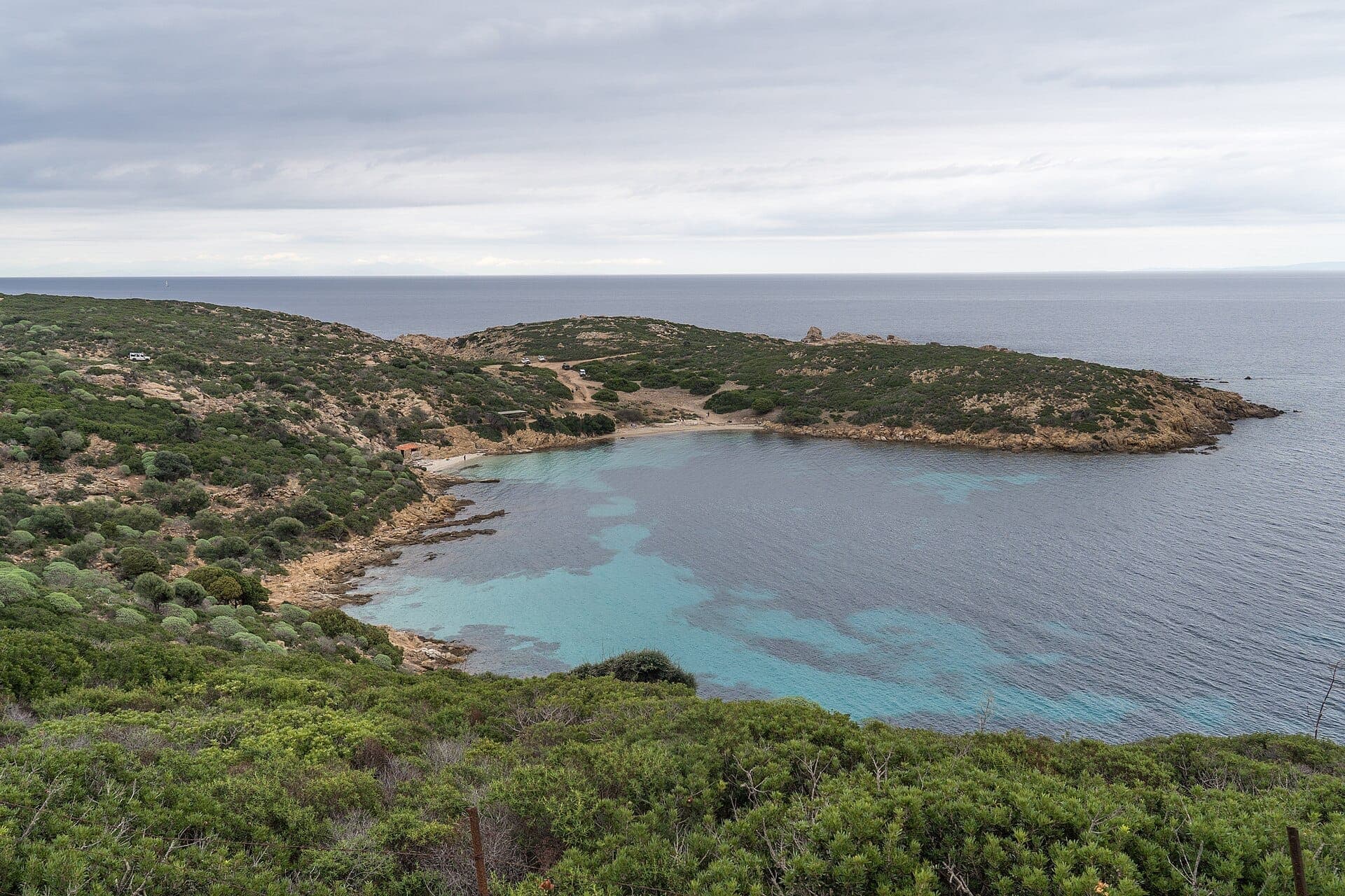 Eine abgelegene Bucht im Parco Nazionale dell'Asinara mit türkisfarbenem Wasser, felsiger Küste und üppigen grünen Hügeln unter einem bewölkten Himmel.