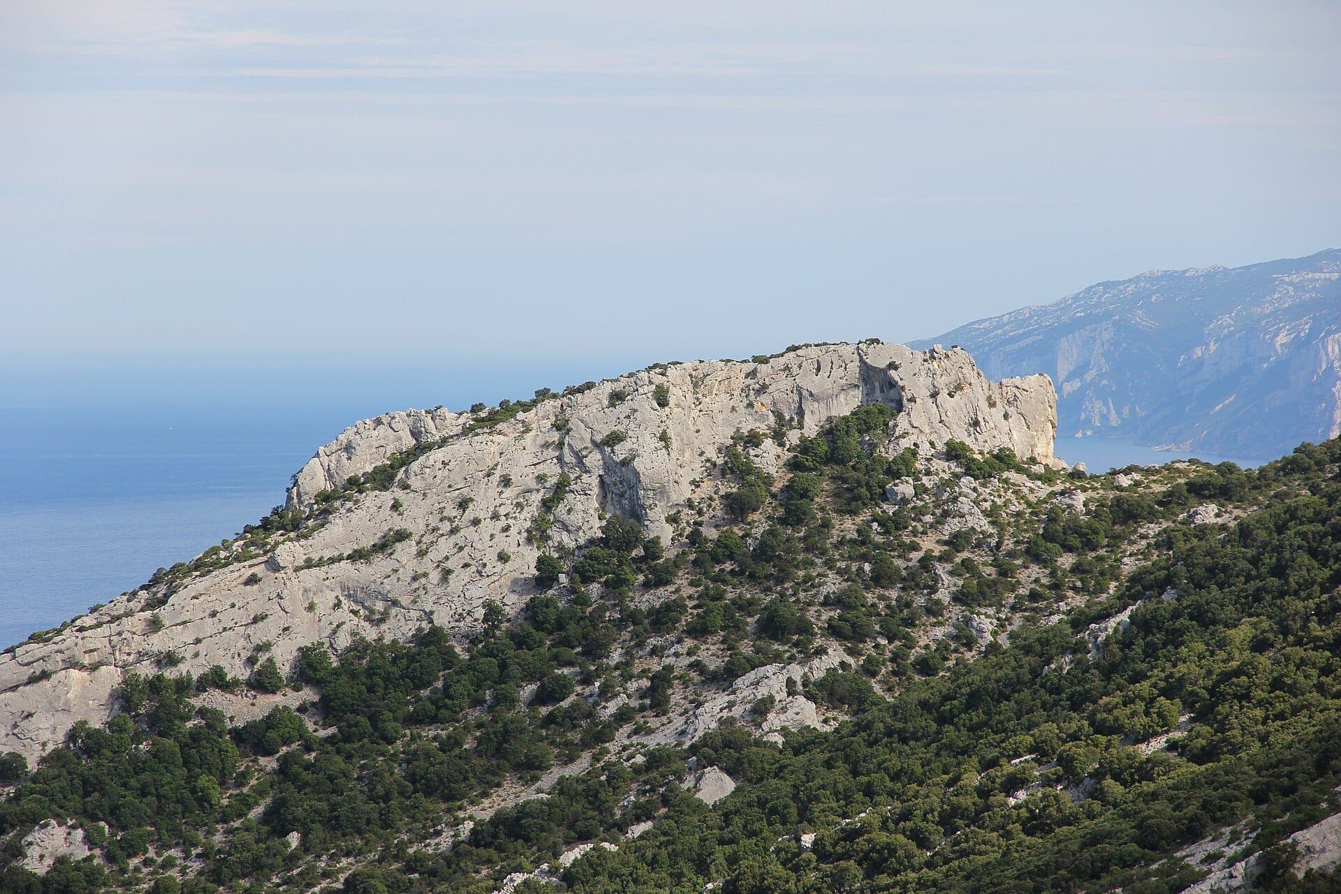 Acantilados rocosos de piedra caliza y vegetación verde del Parco Nazionale del Golfo di Orosei e del Gennargentu, con vistas lejanas del mar azul y las montañas.