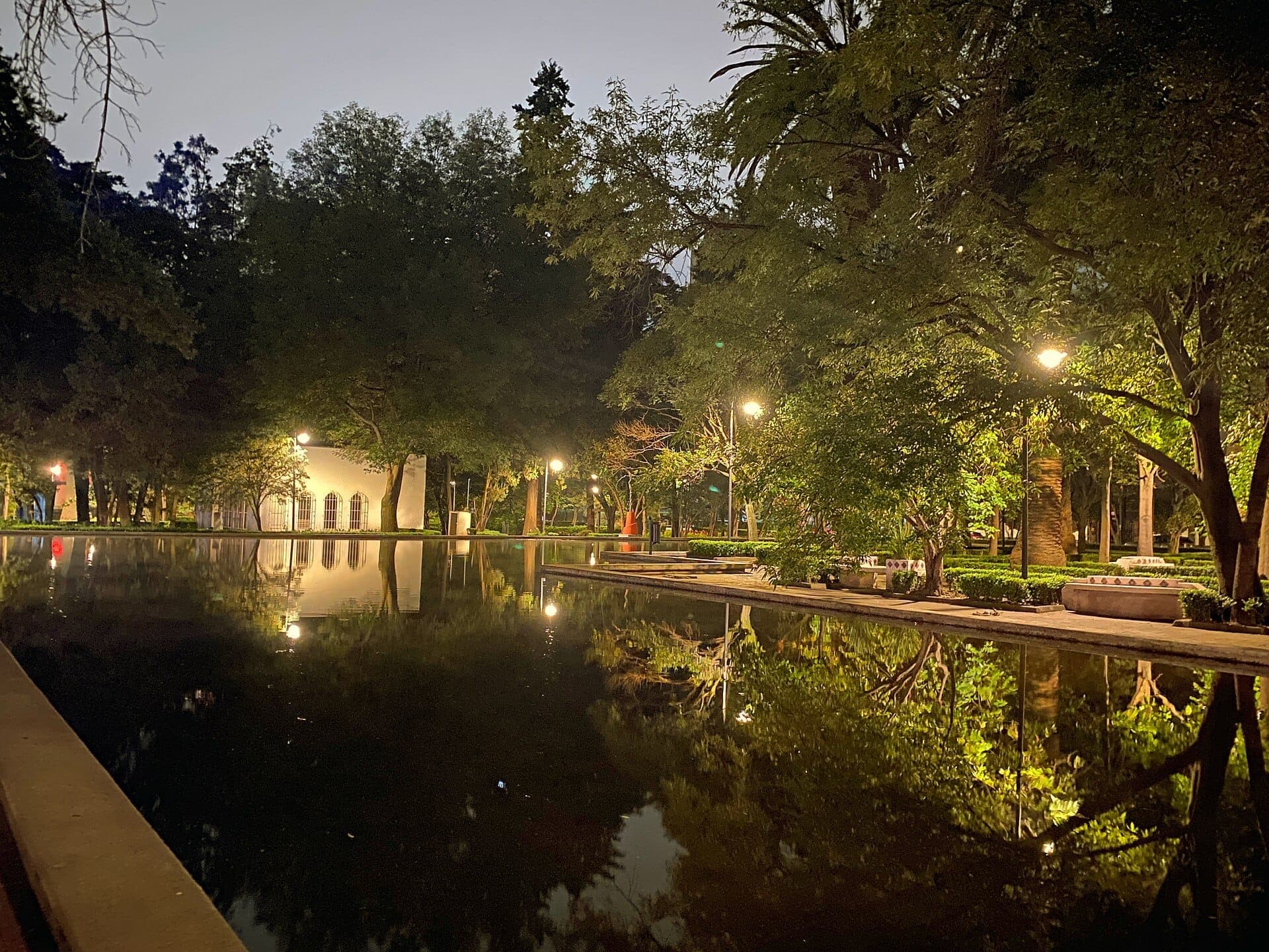 สระสะท้อนแสงที่ล้อมรอบด้วยต้นไม้เขียวขจีและม้านั่งในสวนยามค่ำคืนที่ Parque Lincoln พร้อมแสงไฟถนนอบอุ่นส่องสว่างบรรยากาศอันสงบ