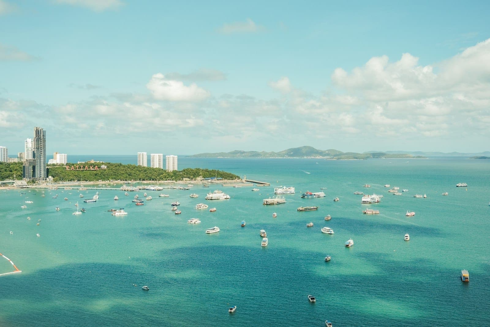 ภาพถ่ายทางอากาศของอ่าวพัทยาที่มีเรือกระจายอยู่บนน้ำสีเขียวอมฟ้า พร้อมเส้นขอบฟ้าของเมืองและเนินเขาสีเขียวในระยะไกลใต้ท้องฟ้าที่มีเมฆบางส่วน