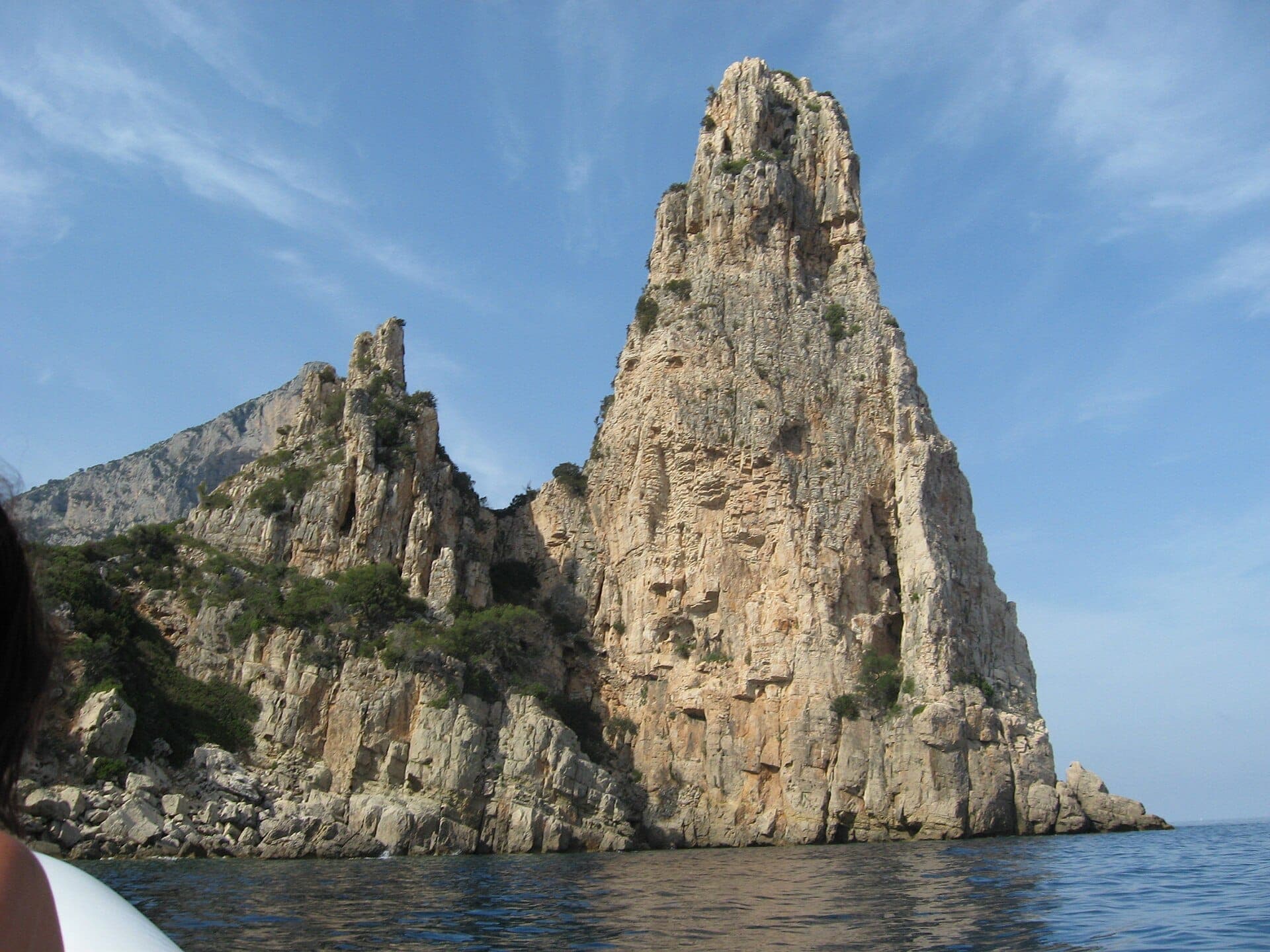 El imponente pináculo de piedra caliza de Pedra Longa emerge directamente del claro mar Tirreno azul, con escarpados acantilados y escasa vegetación bajo un cielo soleado.
