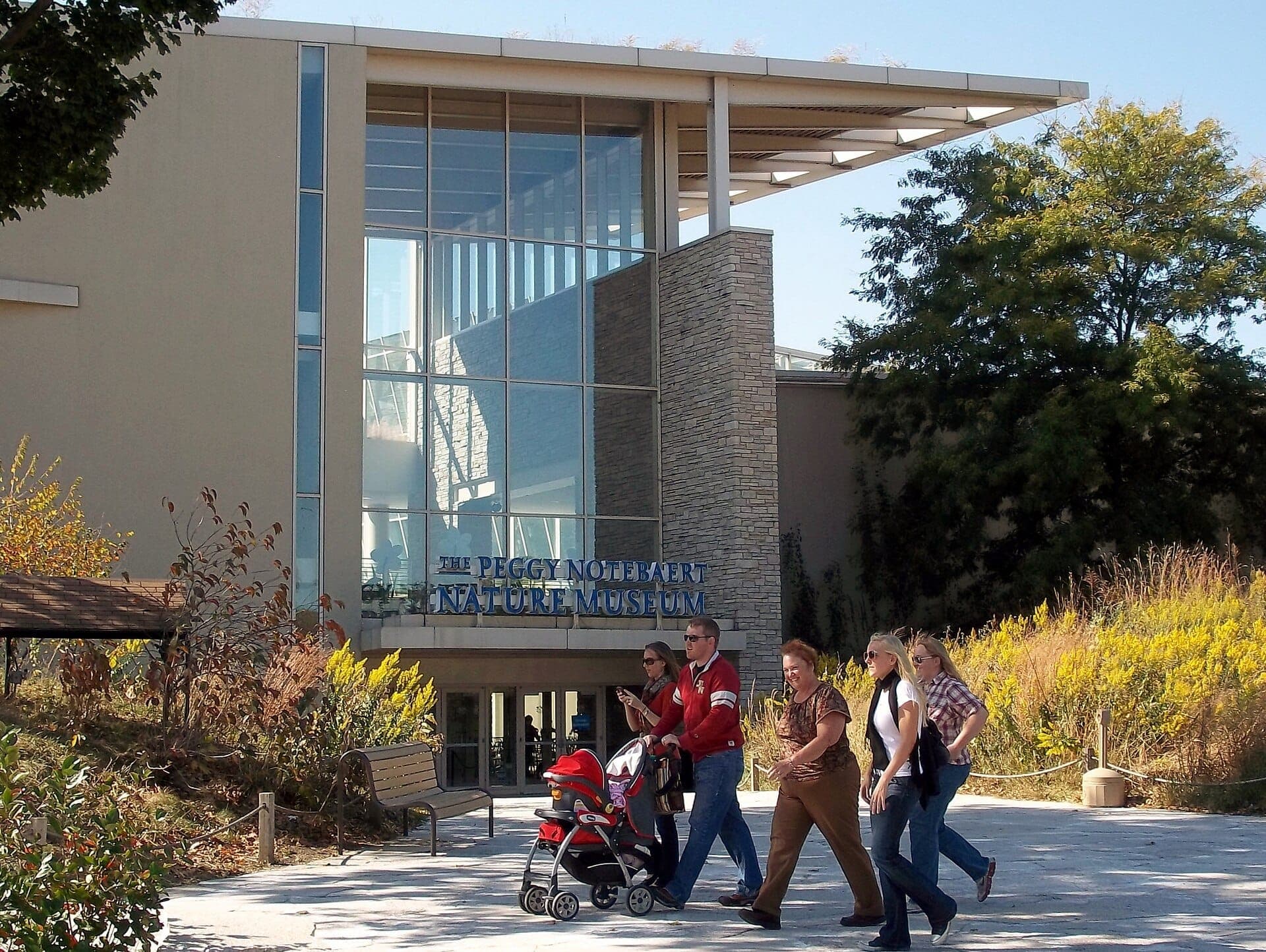 Glass-front entrance of the Peggy Notebaert Nature Museum with modern architecture, lush landscaping, and a family group walking on a sunny day.