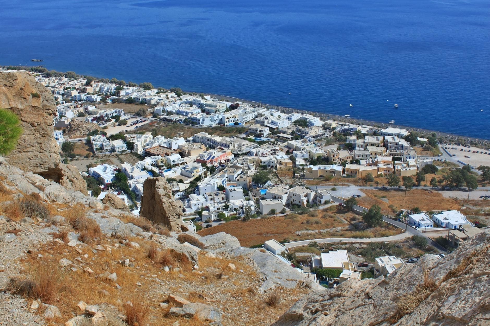 Luftaufnahme des Dorfes Perissa auf Santorini, mit weiß getünchten Gebäuden, trockenen Felshängen und dem tiefblauen Ägäischen Meer jenseits des Schwarzsandstrandes.
