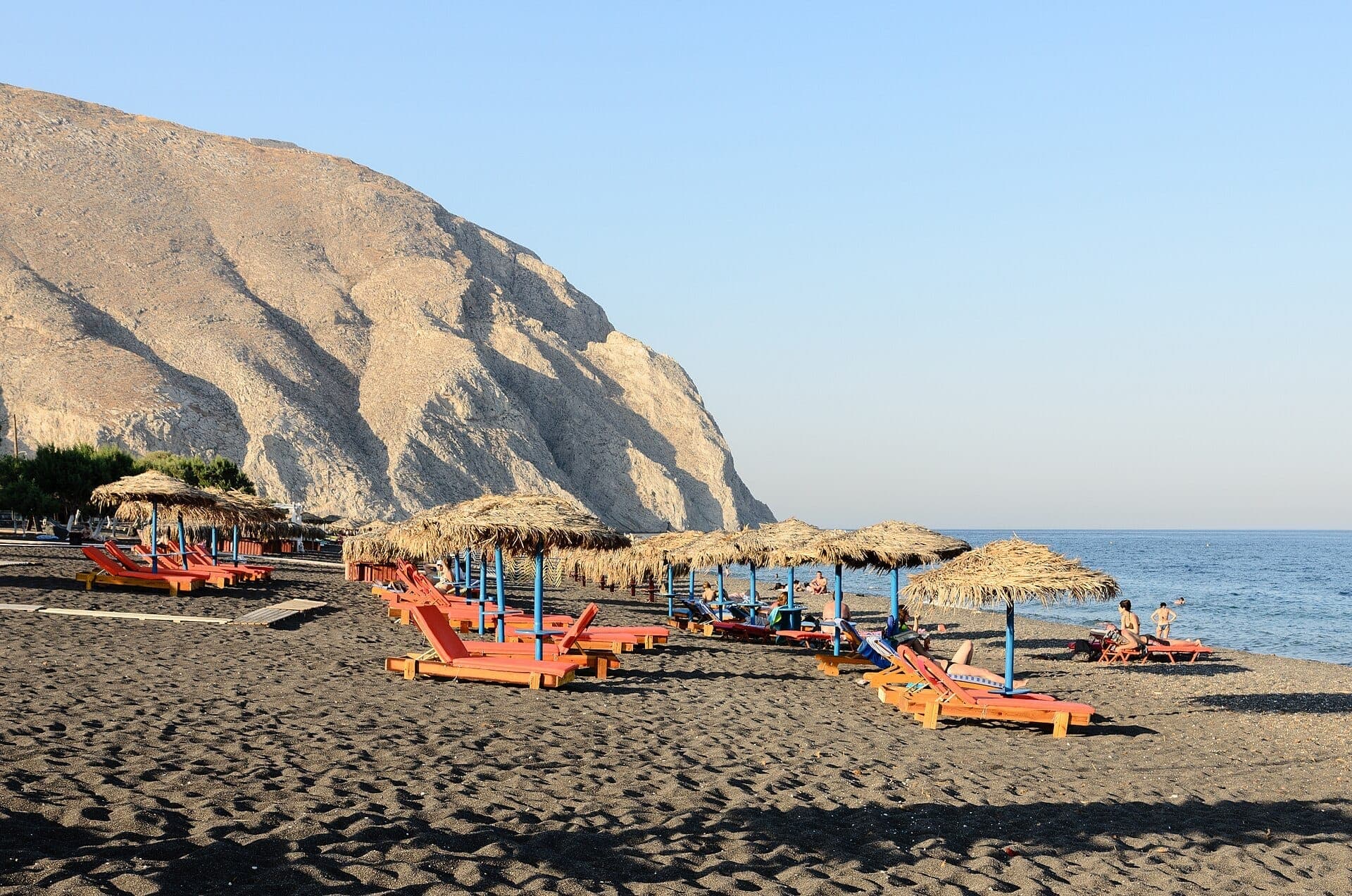 Reihen von Liegestühlen und Strohschirmen stehen auf dem schwarzen Vulkansand des Strandes Perissa, mit dem imposanten Mesa Vouno im Hintergrund.