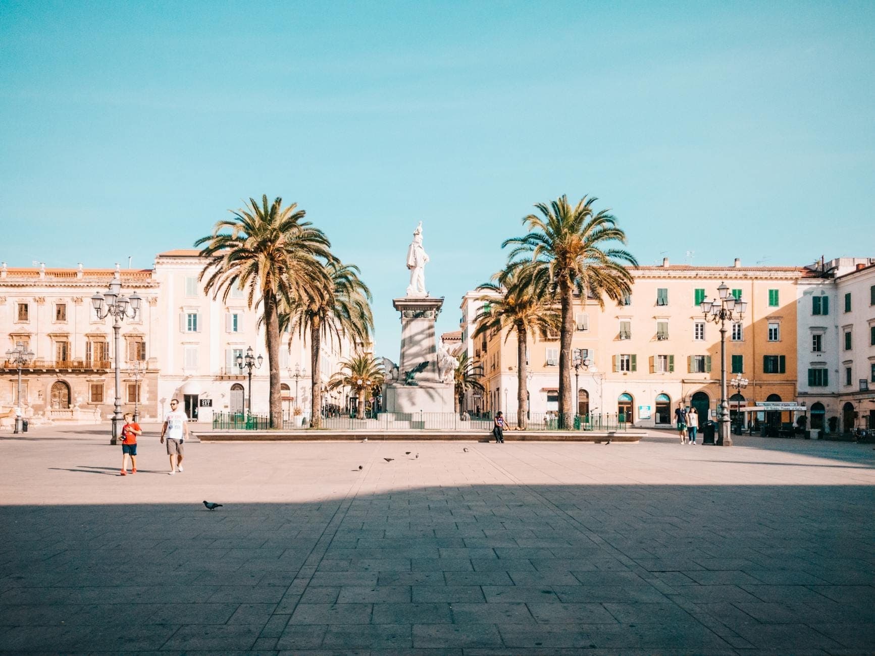 Vista panorámica de la Piazza d'Italia en Sassari con edificios neoclásicos, palmeras y una estatua real del siglo XIX en el centro bajo un cielo azul despejado.
