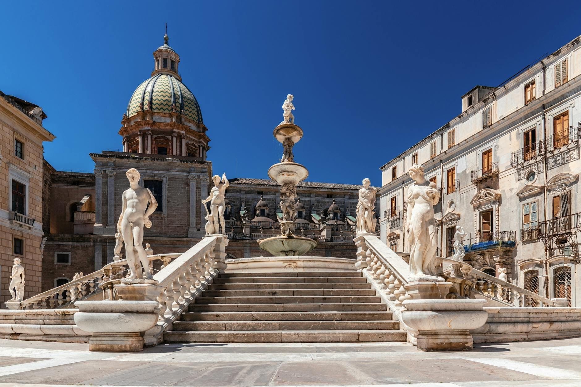Pemandangan luas tangga megah Piazza Pretoria dan air mancur Renaisans yang dikelilingi patung-patung telanjang bertema mitologi, dengan bangunan bersejarah dan langit biru cerah di Palermo, Sisilia.