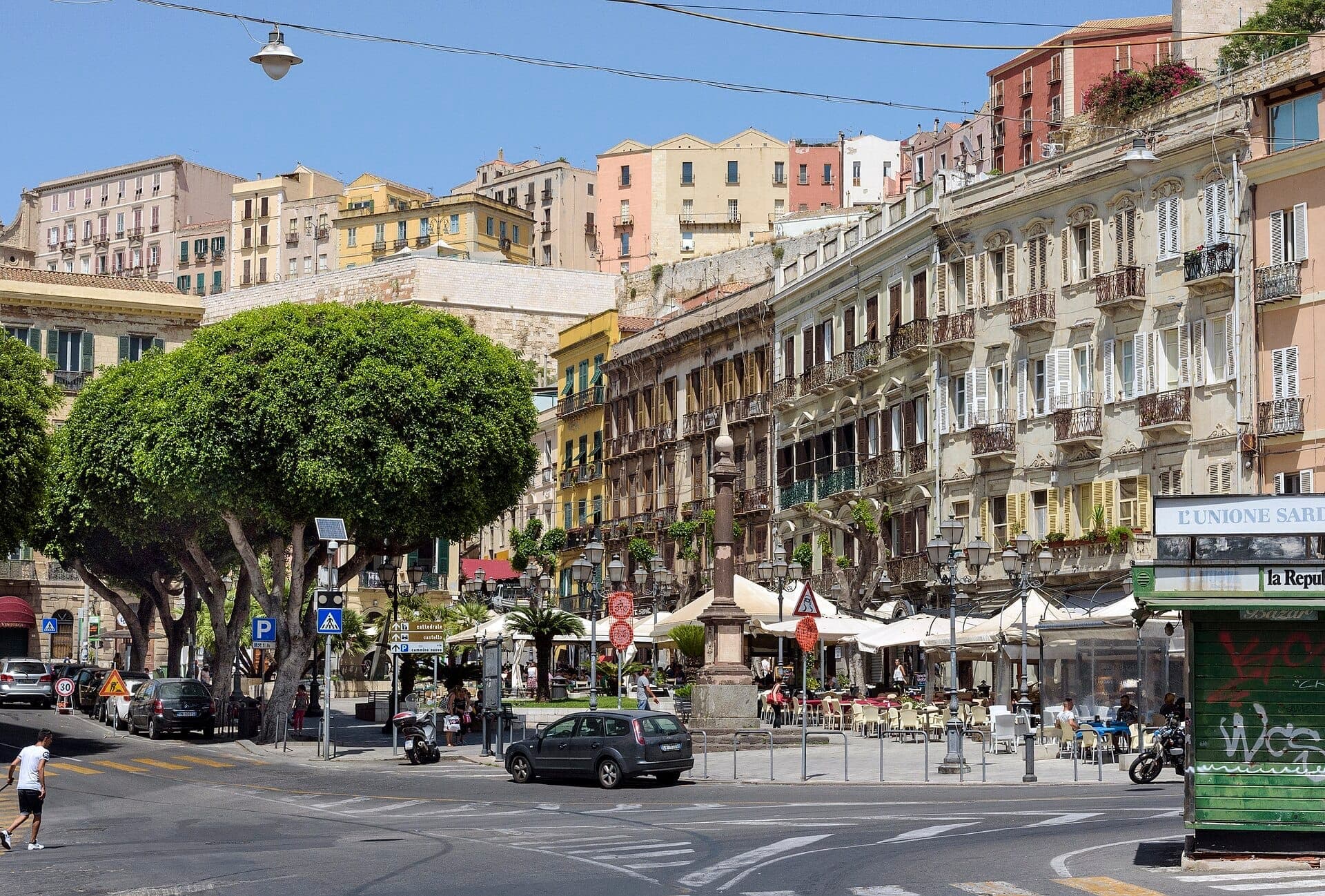 Piazza Yenne en Cagliari con edificios históricos, árboles frondosos, señales de tráfico, cafés al aire libre y una estatua en el centro de la plaza.