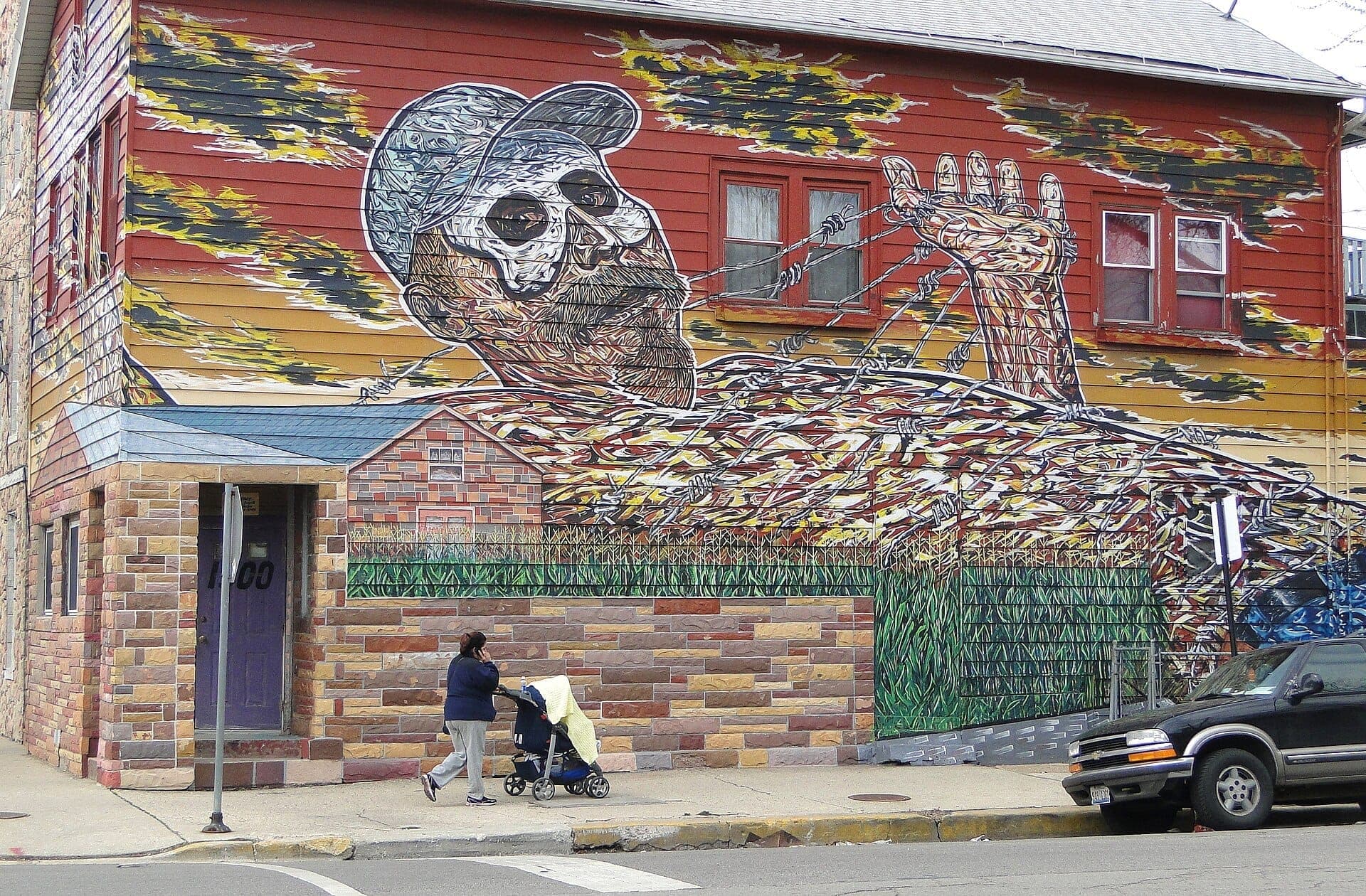 Colorful street mural featuring a man entangled in barbed wire on a building in Chicago's Pilsen neighborhood, with a person pushing a stroller nearby.