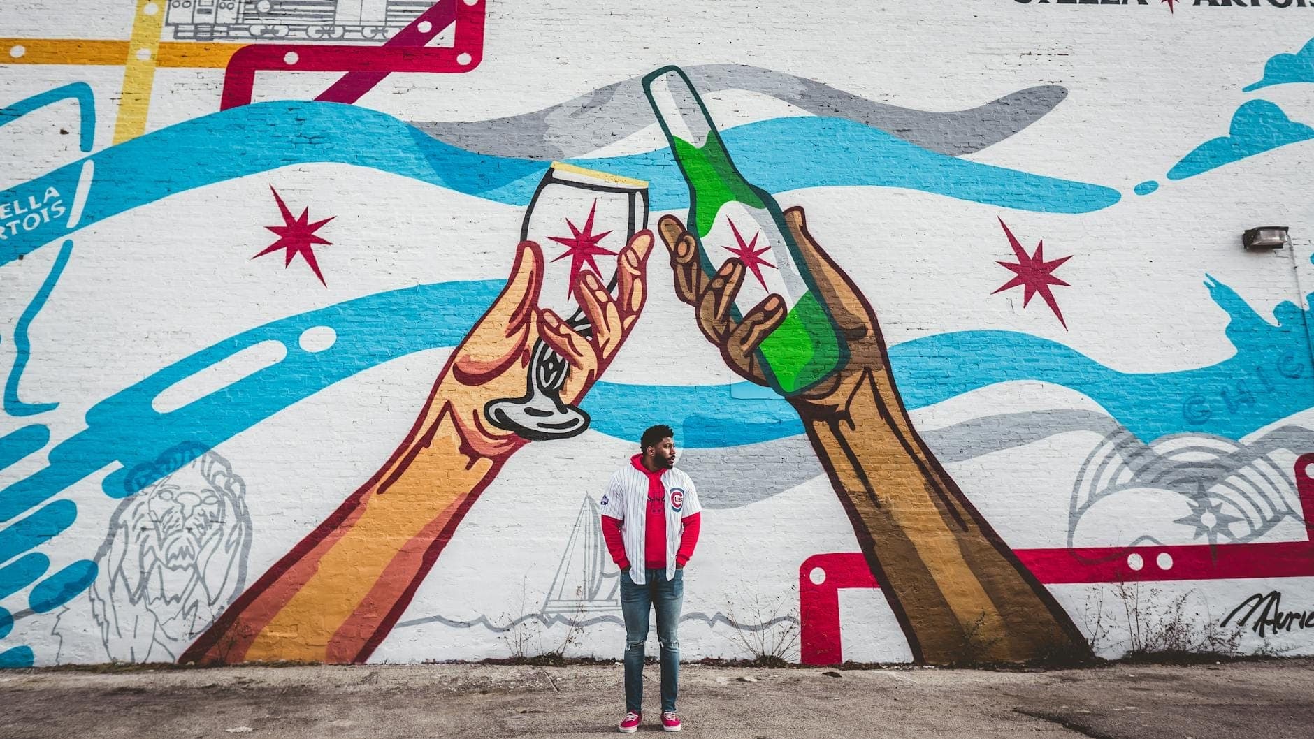 A colorful mural on a wall featuring two large hands toasting with a beer bottle and wine glass, with a person standing in front. Chicago-style stars and waves complete the vibrant street art scene.