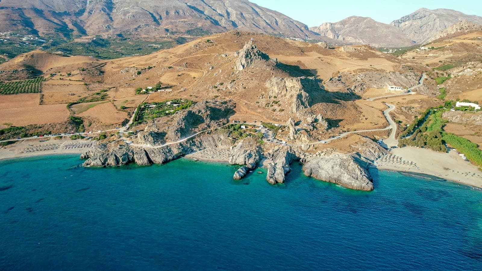 Weitläufige Luftaufnahme des Plakias Beach an Kretas Südküste mit türkisfarbenem Wasser, sandigem Halbmond-Ufer, Liegestühlen und schroffen Bergen im Hintergrund.