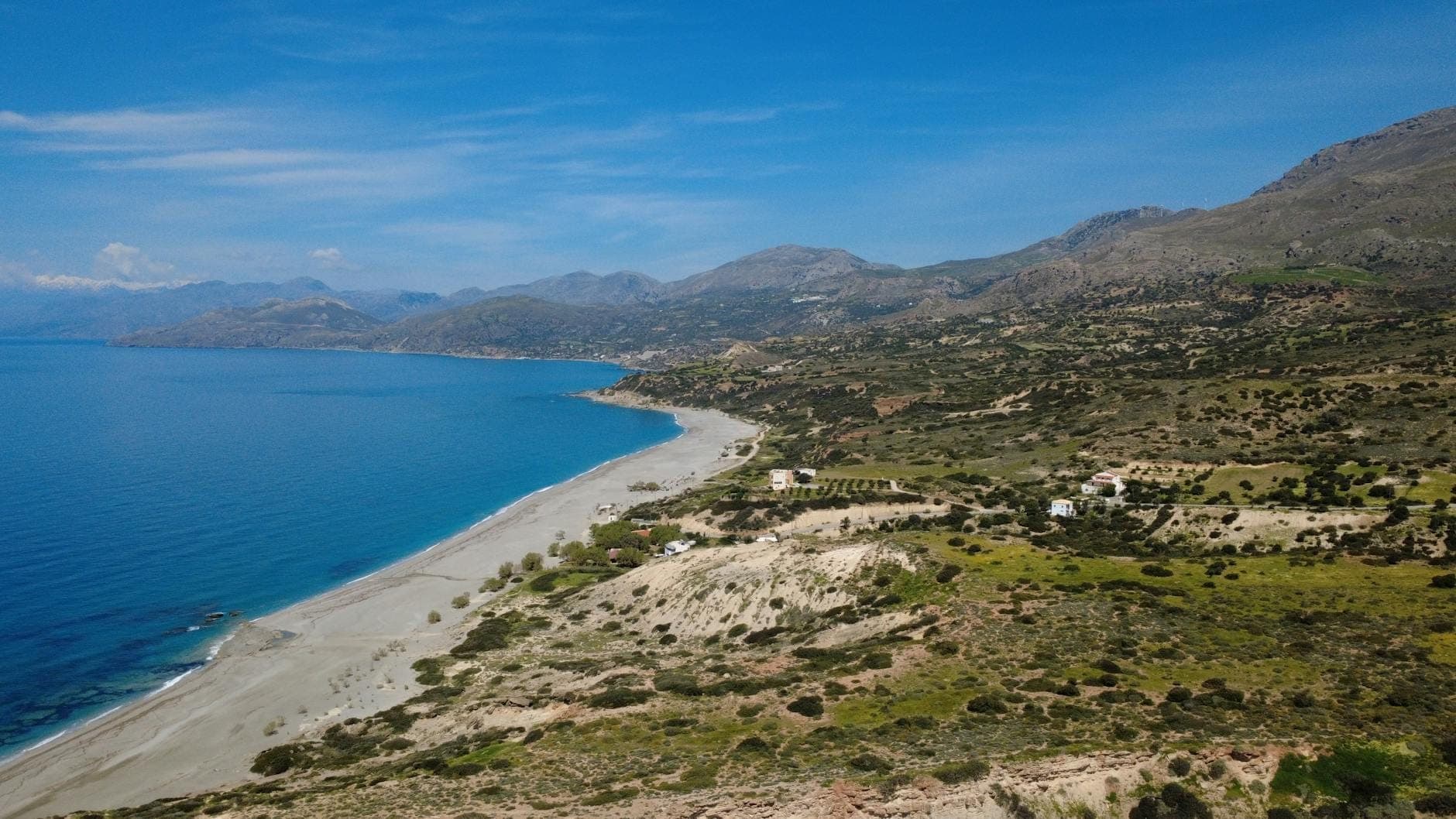 Ein breiter, geschwungener Sandstrand an der Südküste Kretas, eingerahmt von grünen Hügeln und Bergen unter einem strahlend blauen Himmel.