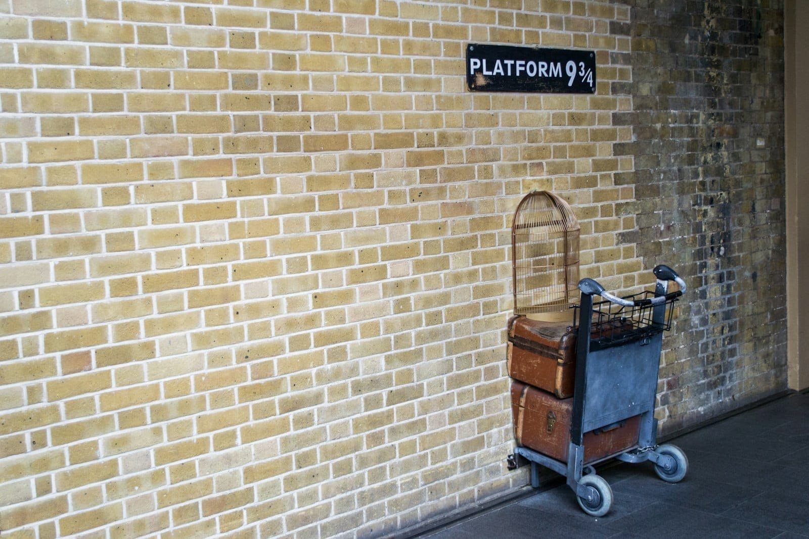 Ein Gepäckwagen mit Vintage-Koffern und einem Vogelkäfig steckt halb in der Backsteinwand unter dem Schild zu Gleis 9¾ am King's Cross.