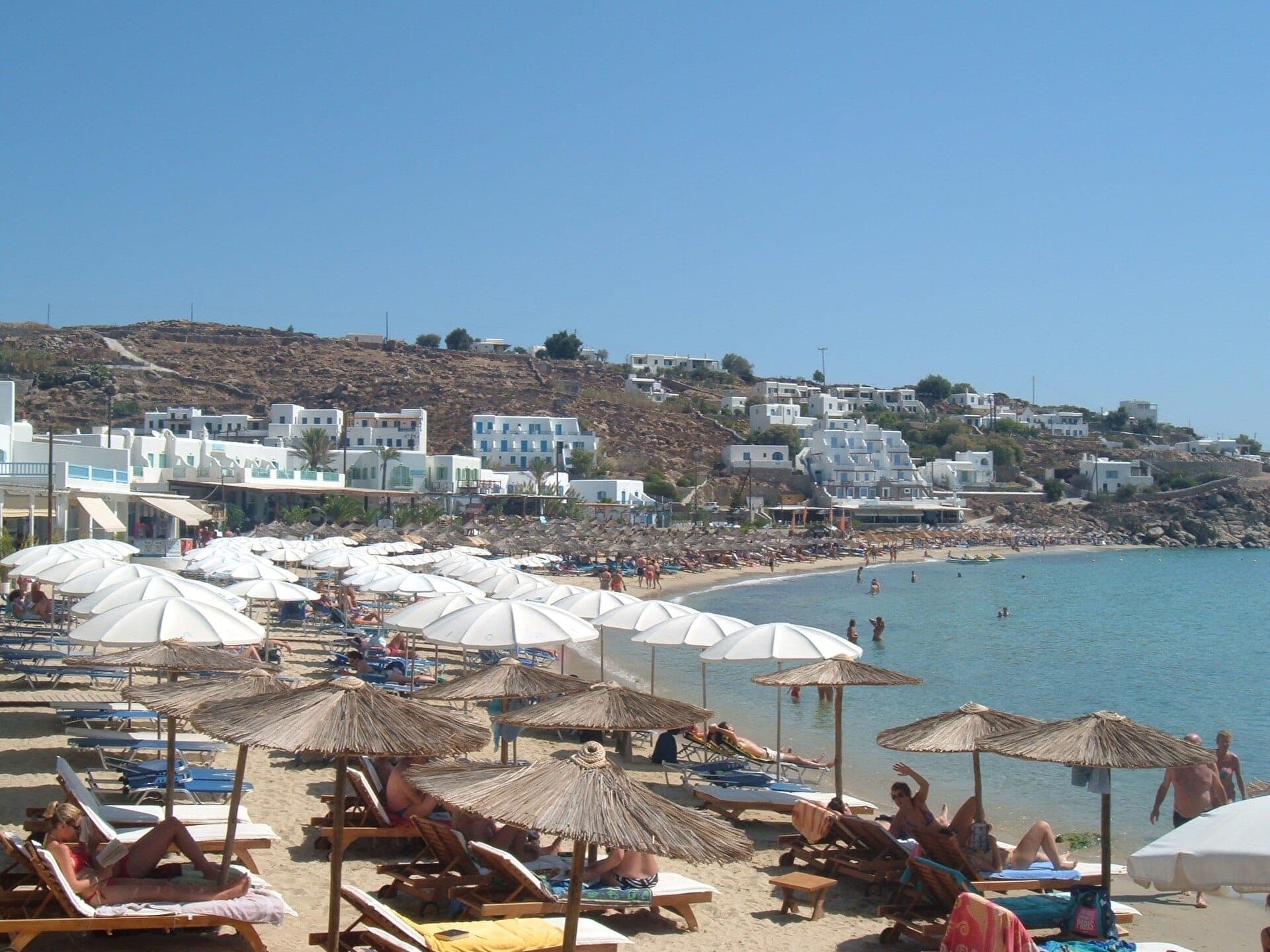 Der Strand Platis Gialos mit Reihen von Sonnenliegen und Sonnenschirmen auf goldenem Sand, klarem blauem Wasser und weiß getünchten Hotels am Hang in Mykonos.