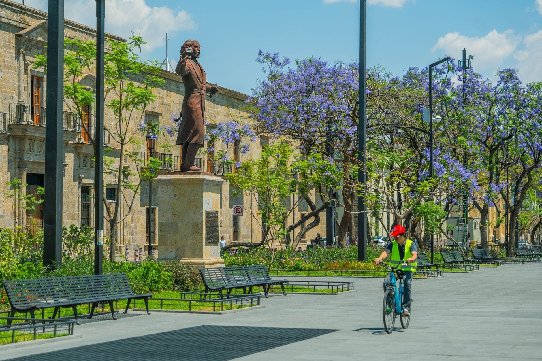 วิวของ Plaza de la Liberación มีรูปปั้น ม้านั่ง ต้นศรีตรังสีม่วงกำลังบาน และนักปั่นจักรยาน ผ่านจัตุรัสคนเดินกลางย่านเมืองเก่ากัวดาลาฮารา