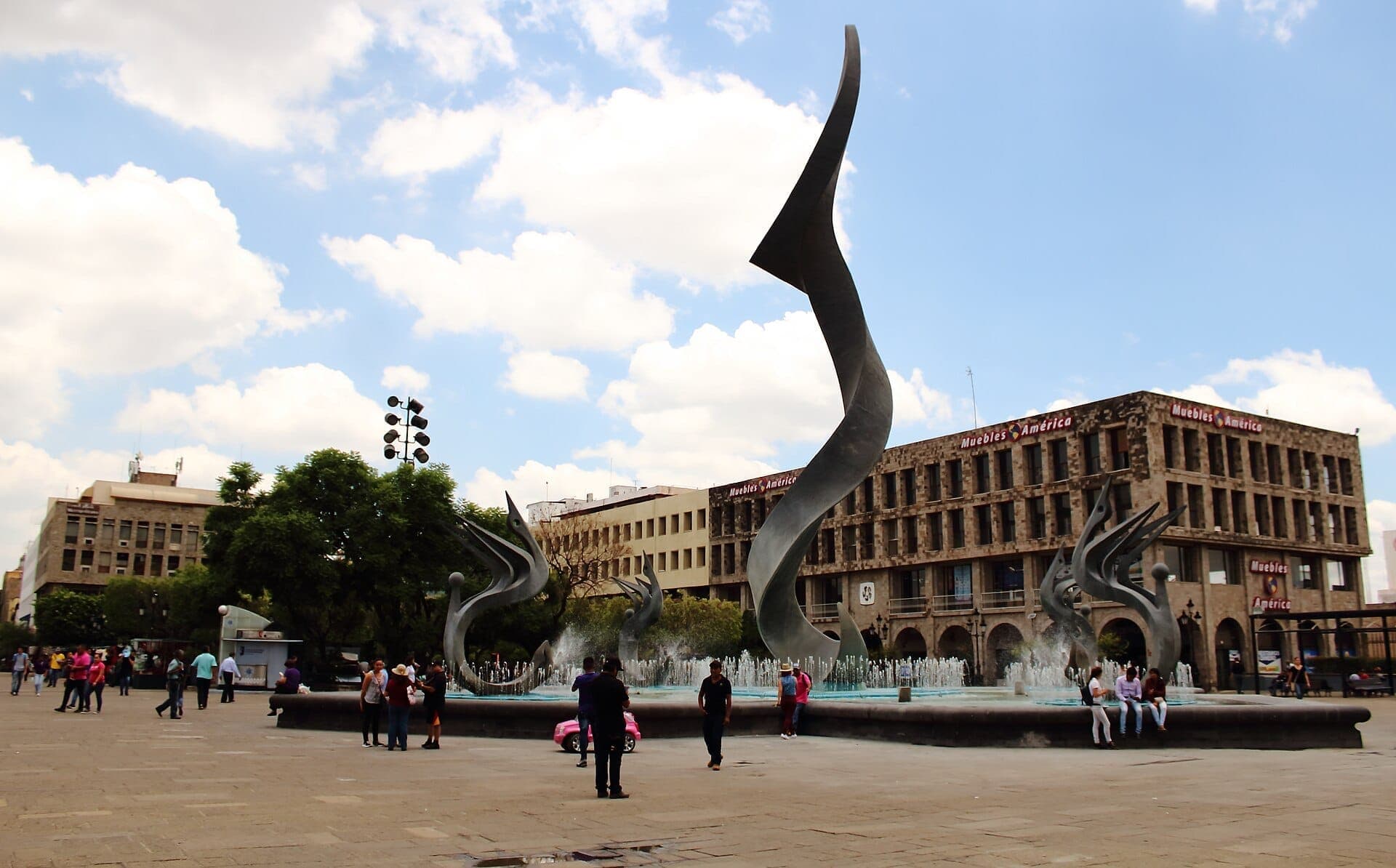 วิวกว้างของ Plaza Tapatía ที่มีประติมากรรมโลหะสมัยใหม่ น้ำพุขนาดใหญ่ อาคารราชการโดยรอบ และผู้คนที่เดินเล่นใต้ท้องฟ้าที่มีเมฆบางส่วน