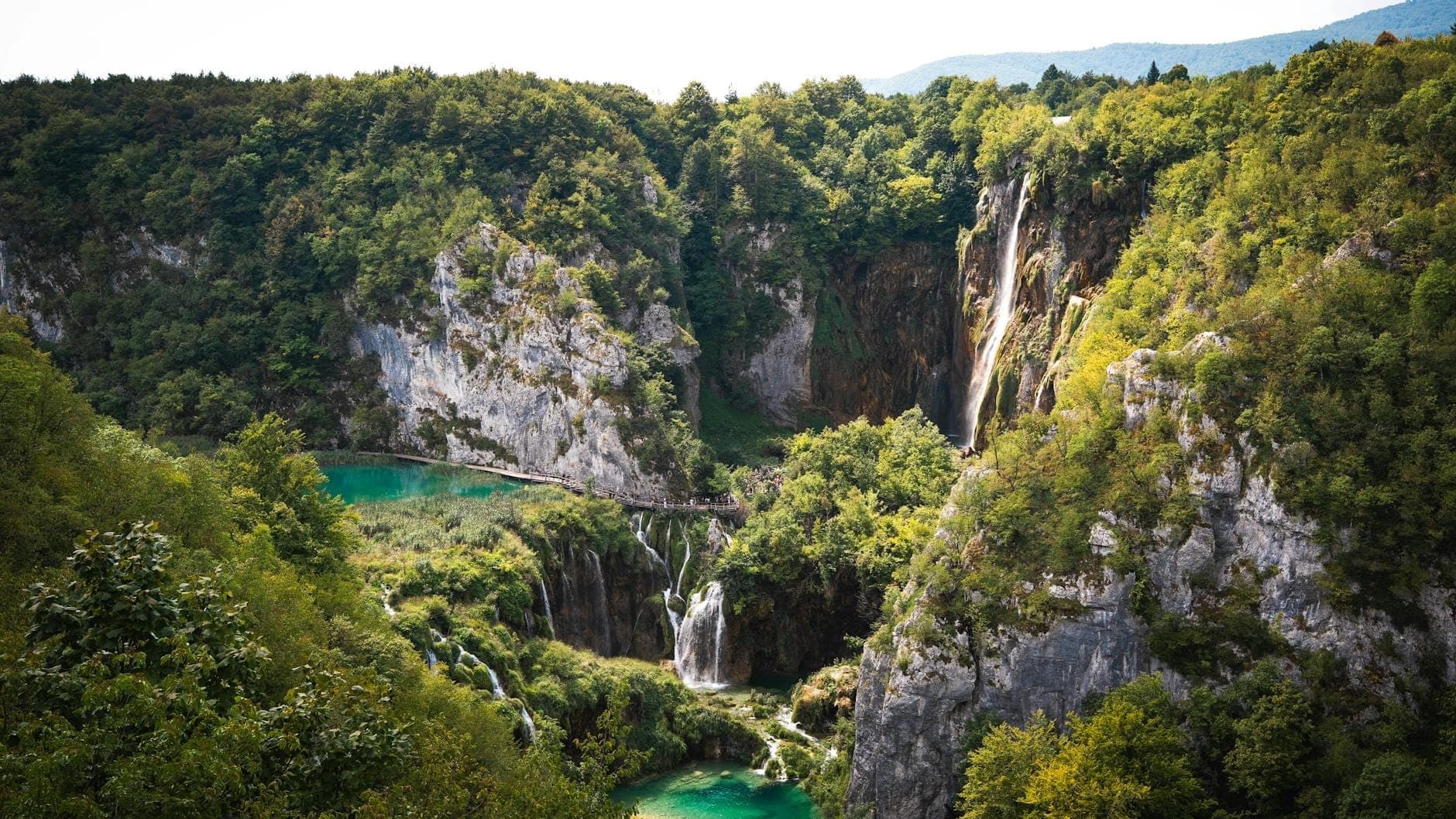 Vue panoramique du parc national des lacs de Plitvice avec ses multiples lacs turquoise, ses chutes d'eau en cascade, ses falaises calcaires et sa forêt luxuriante sous un ciel lumineux.