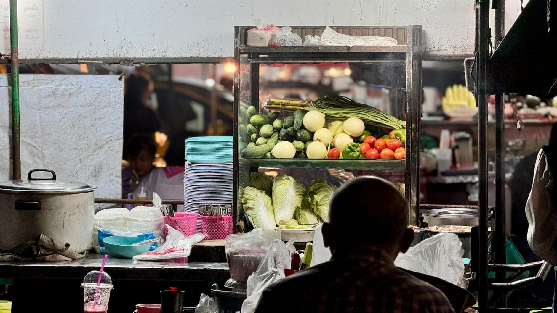 Marktstand mit hell erleuchteten frischen Gemüsesorten und Küchenutensilien, von hinten durch einen sitzenden Gast in einer entspannten und lebhaften Abendatmosphäre fotografiert.