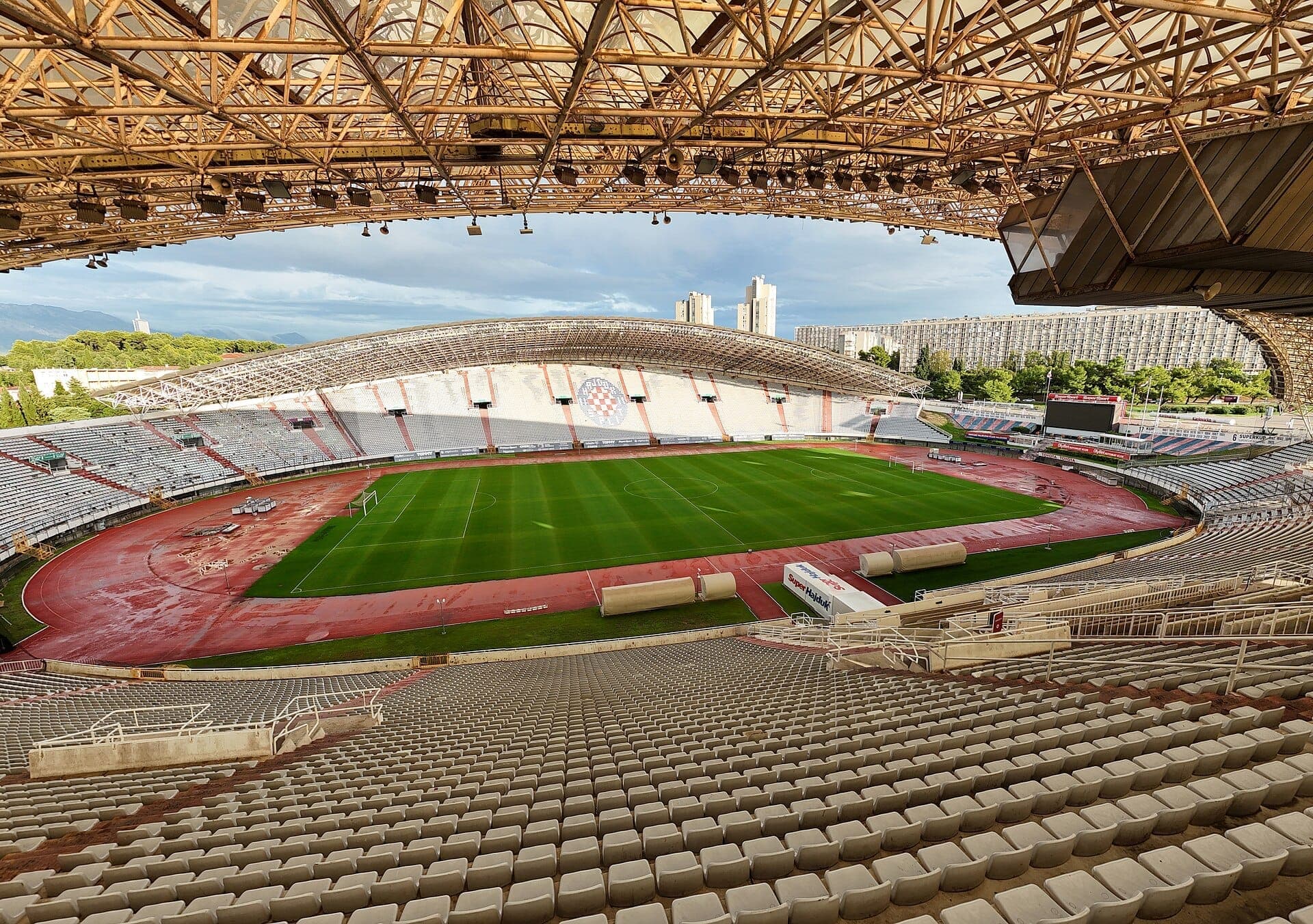 Vista interna ampia dello Stadio Poljud che mostra il suo iconico tetto a forma di conchiglia, tribune vuote, campo da calcio verde e lo skyline urbano circostante a Split.